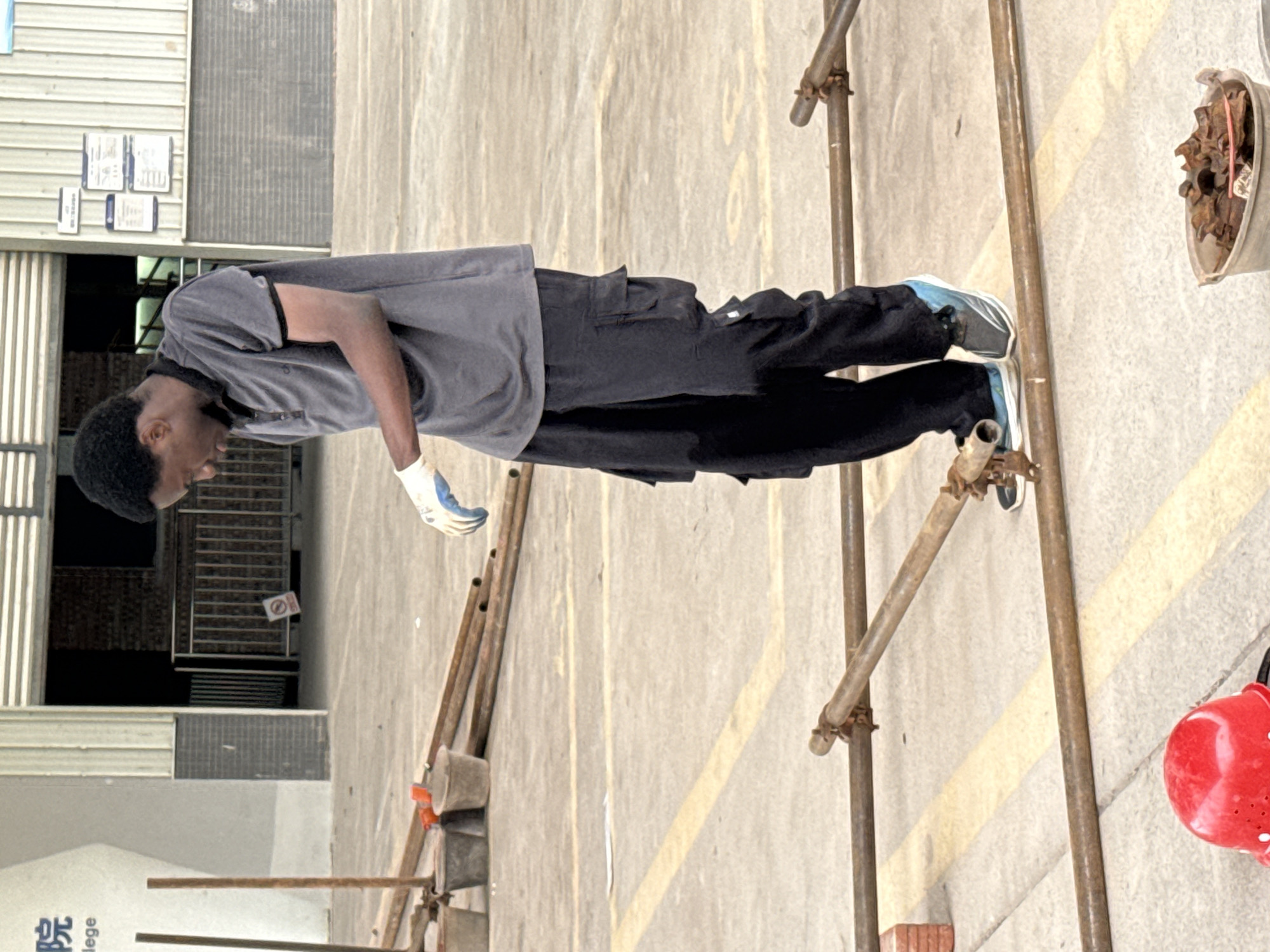A young Black man wearing a grey polo shirt, black cargo pants, white and blue athletic shoes, and white work gloves stands on a concrete surface. He is looking down and appears to be working with scaffolding. The scaffolding consists of rusty metal pipes that are joined together. In the background, there is a building with signage that includes Chinese characters and the word "lege." There are also metal barriers and what appear to be information boards on the building. In the foreground, there are several buckets and a red hard hat. The overall environment suggests an outdoor work or construction site.