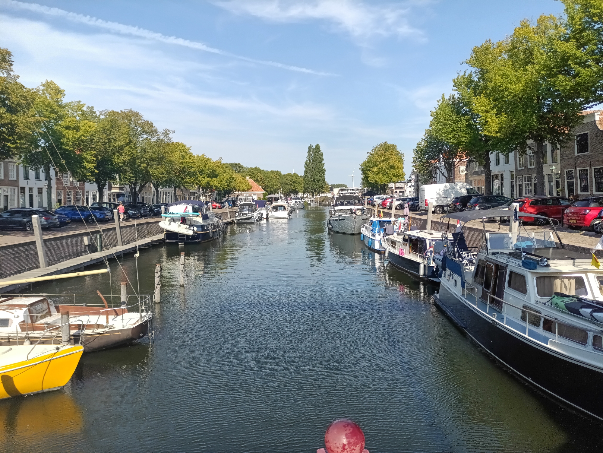 This is a daytime, eye-level, wide shot of a canal in the city of Brielle, Netherlands. The canal runs horizontally across the frame, with boats docked on both sides. The water is calm and reflects the blue sky and surrounding trees.
On the left bank of the canal, there is a row of parked cars. Behind the cars, there are several multi-story buildings with traditional Dutch architecture, featuring brick facades and white-trimmed windows. A line of lush green trees with full canopies separates the buildings from the canal. Several boats are docked along this side, including a yellow sailboat in the foreground.
On the right bank, more boats are docked along a paved walkway. These boats appear to be houseboats or motor yachts. Behind them are more buildings, similar in style to those on the left, with trees interspersed along the waterfront.
In the distance, the canal continues to curve, with more boats and buildings visible. The sky is clear blue with scattered white clouds. The overall scene depicts a peaceful, urban waterway lined with residential and recreational vessels, characteristic of a Dutch canal town. There are no visible people actively engaged in any actions within the frame.