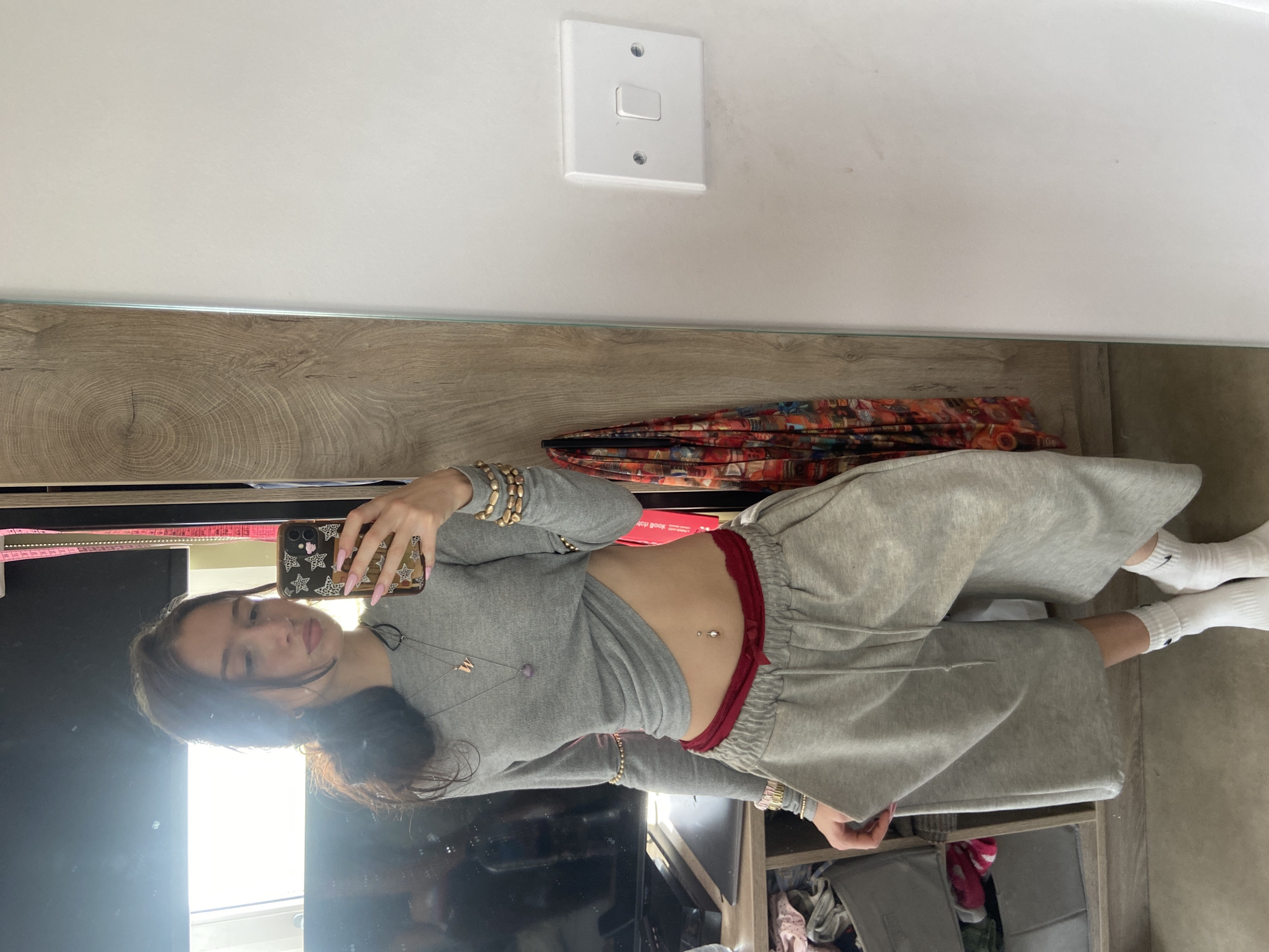 A woman stands in front of a mirror, taking a selfie with her phone. She is wearing a grey cropped long-sleeved shirt and grey sweatpants with a red drawstring. She also has white socks. The background includes a wall with a light switch, a wooden trim, a shelf with storage bins, and other items. The location is unspecified and cannot be determined from the provided image.