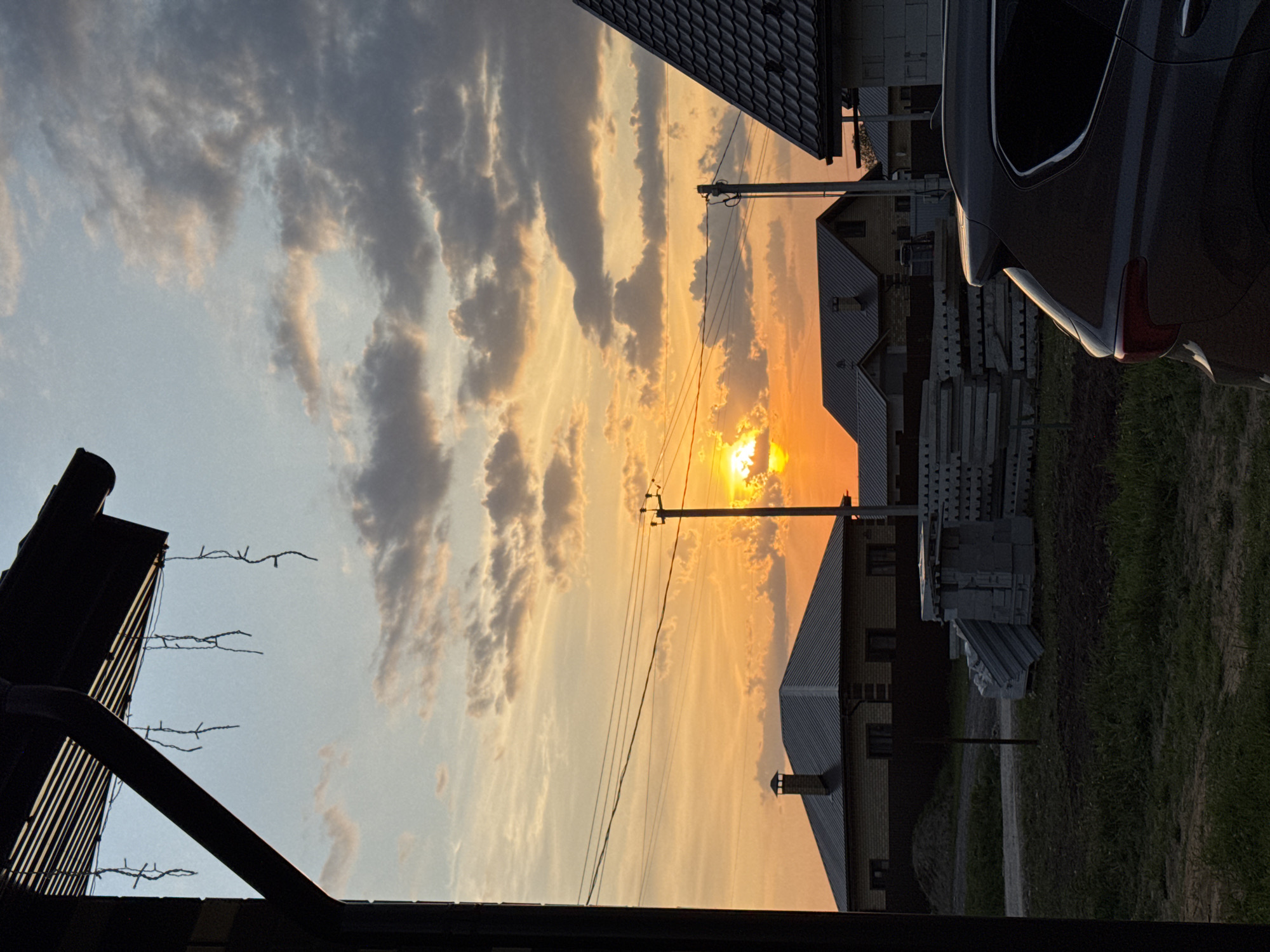The image depicts a sunset over the town of Ферма, Russia. The sky is filled with orange and grey clouds, with the sun visible between the clouds. A power line runs horizontally across the image. In the foreground, there are houses with grey roofs and a car is parked. Construction materials are present near the building. The foreground is partially obscured by a dark structure, possibly part of a building.
