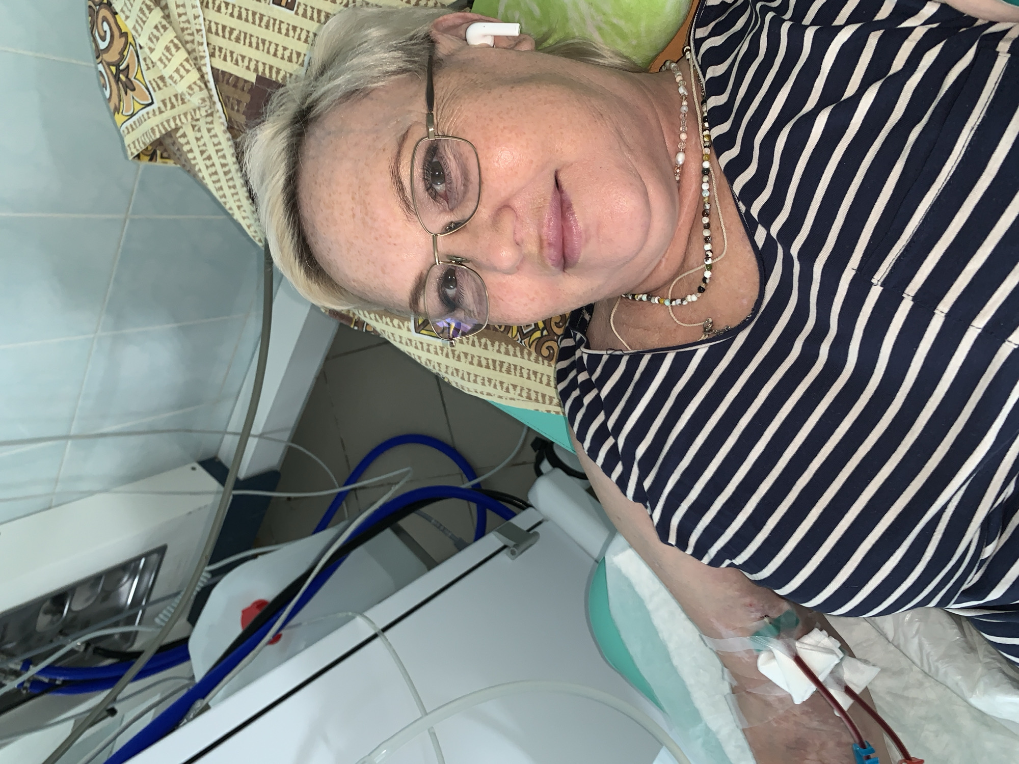 A middle-aged woman with short, light blonde hair and glasses is lying down, possibly in a medical facility in Chaikovsky, Russia. She is wearing a striped shirt and has an earbud in her ear. Medical tubing is connected to her arm, with visible blood. The surrounding environment includes medical equipment with blue tubes and a patterned cushion. The scene suggests a medical procedure, possibly dialysis, is taking place.