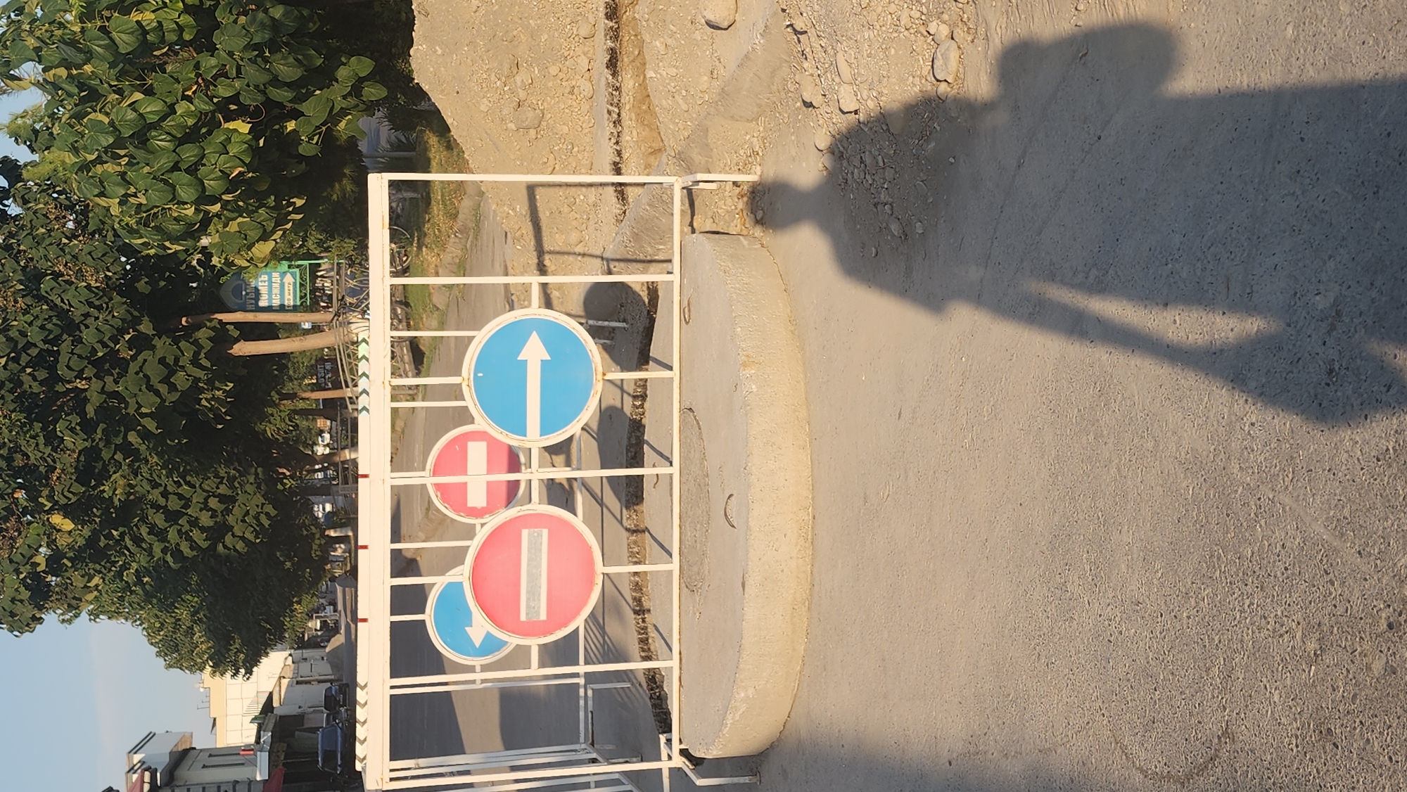 In the city of Unknown, Unknown, on a paved road, a white metal barrier is erected. Attached to the barrier are four circular traffic signs: two "Do Not Enter" signs and two "Turn Right" signs, with the "Do Not Enter" signs on the left. In front of the barrier is a concrete ring, likely part of roadwork. The road is under construction, with piles of dirt and rocks visible on the right side of the image. A shadow of a person, holding a bag, is cast on the road in the foreground. Trees and buildings line the street in the background. The sky is clear and blue.