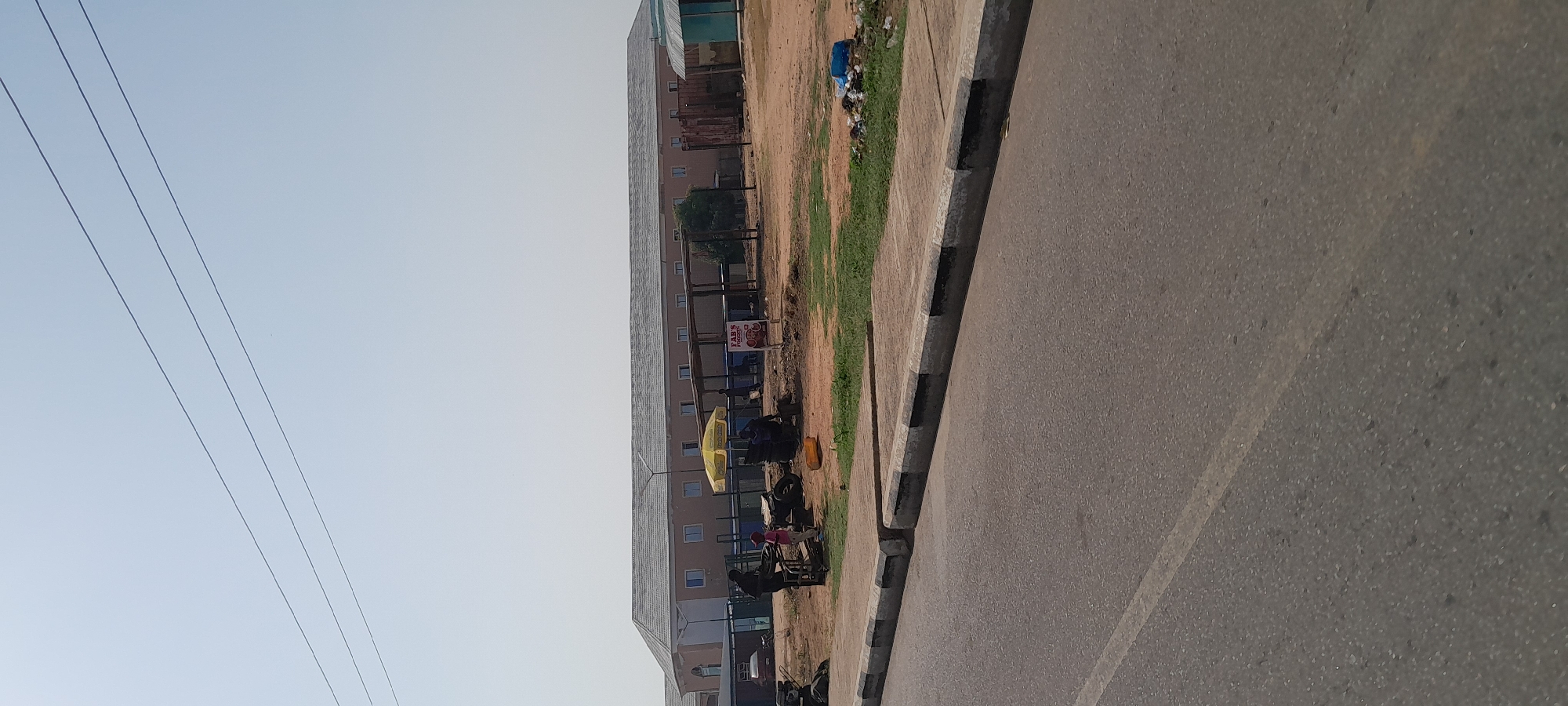 A street-level view of a commercial area in Jalingo, Nigeria, under a clear sky. In the background, a long, beige-colored building with a gray, tiled roof stretches horizontally, featuring numerous evenly spaced windows. In front of this building, there's an open-air market area with several stalls and structures, some with corrugated metal roofing. A yellow umbrella shades a small seating area. In the foreground, a paved road runs diagonally across the lower portion of the frame, bordered by a concrete sidewalk with black and white striped edges. On the sidewalk, a few individuals are visible. To the left, a dark-skinned person stands next to a wheeled cart. A younger person in a pink shirt and dark pants stands near the cart. Further to the right, another individual sits behind a makeshift stall. Tires are stacked near the stall, and various objects, including a yellow jerrycan, are scattered around the area. A sign with text and images, possibly advertising food, is posted near one of the stalls. The ground in front of the market area is unpaved and appears dusty, with patches of dry grass and debris. Several power lines are visible against the sky.