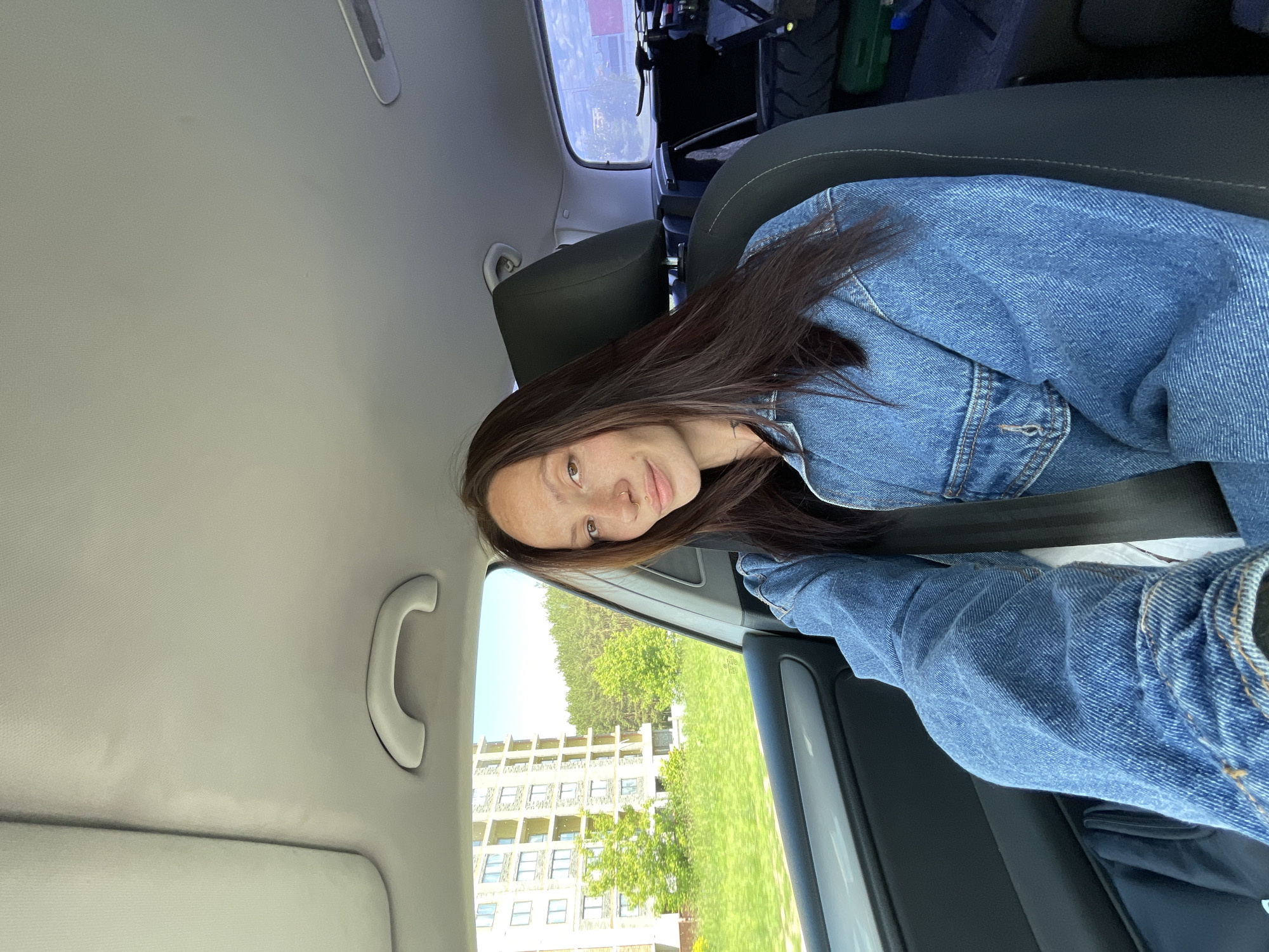 A young woman with long, dark hair and freckles is seated in the passenger seat of a car, wearing a denim jacket. She is looking towards the camera with a slight smile. The car's interior is visible, including the ceiling and a portion of the backseat. Through the window, a tall, modern apartment building and a line of trees are visible against a clear sky, suggesting an urban or suburban setting. The scene appears to be a candid shot taken during a drive.