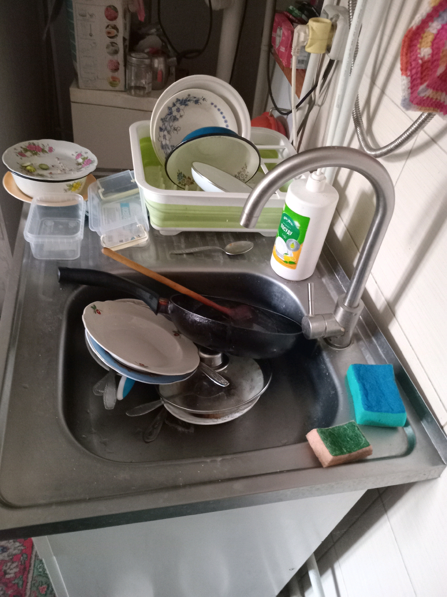 A kitchen sink in Derhachi, Ukraine, is filled with dirty dishes, including plates, bowls, and a frying pan. There are also several plastic containers, a bottle of dish soap, and two sponges on the edge of the sink. The sink and surrounding countertops are made of stainless steel. A faucet with a modern design is visible above the sink. The overall impression is one of a busy kitchen that needs cleaning.