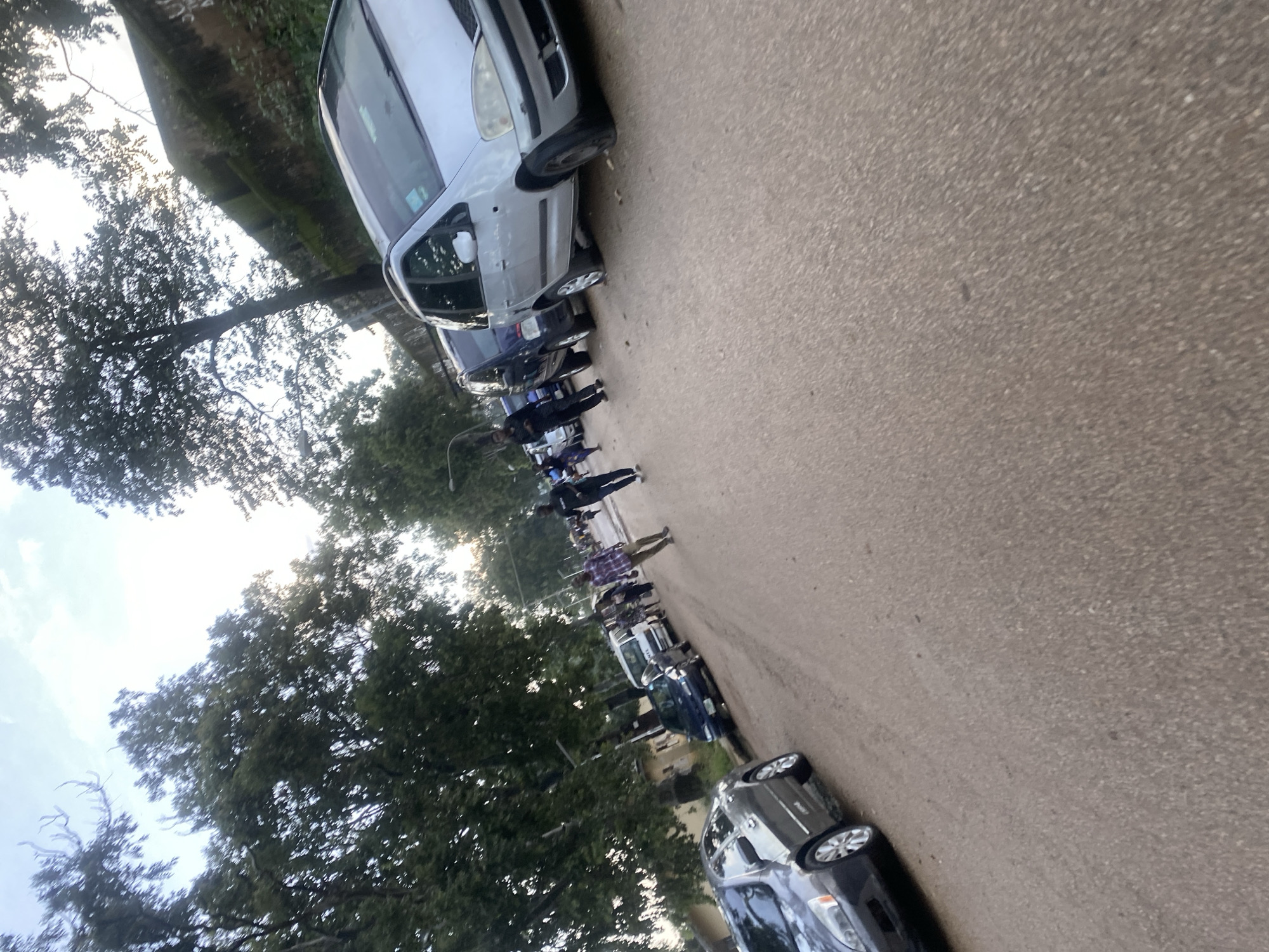 The image captures a street scene in Rafin Par, Nigeria, under a bright, overcast sky with trees lining the road. Several parked cars, including a silver minivan and a dark sedan in the foreground, are visible on the side of the asphalt road. A few individuals are walking along the street, some appearing to be in casual conversation. The environment suggests a typical urban or semi-urban setting in Nigeria, with vegetation and a wall visible in the background.