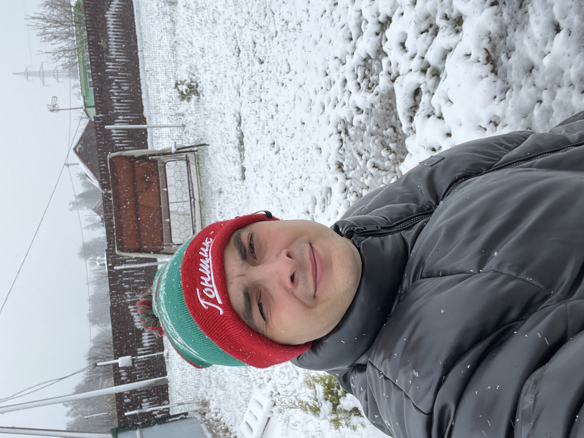 A man wearing a red and green hat with the word "Гонщик" written in white is posing for a selfie outdoors. He is wearing a black puffer jacket with visible snowflakes on it. The background shows a snow-covered yard with a wooden fence and a small building with a red roof. There are power lines visible in the distance, and the scene appears to be in a residential area covered in snow. According to metadata, the location is Раздолье, Russia.