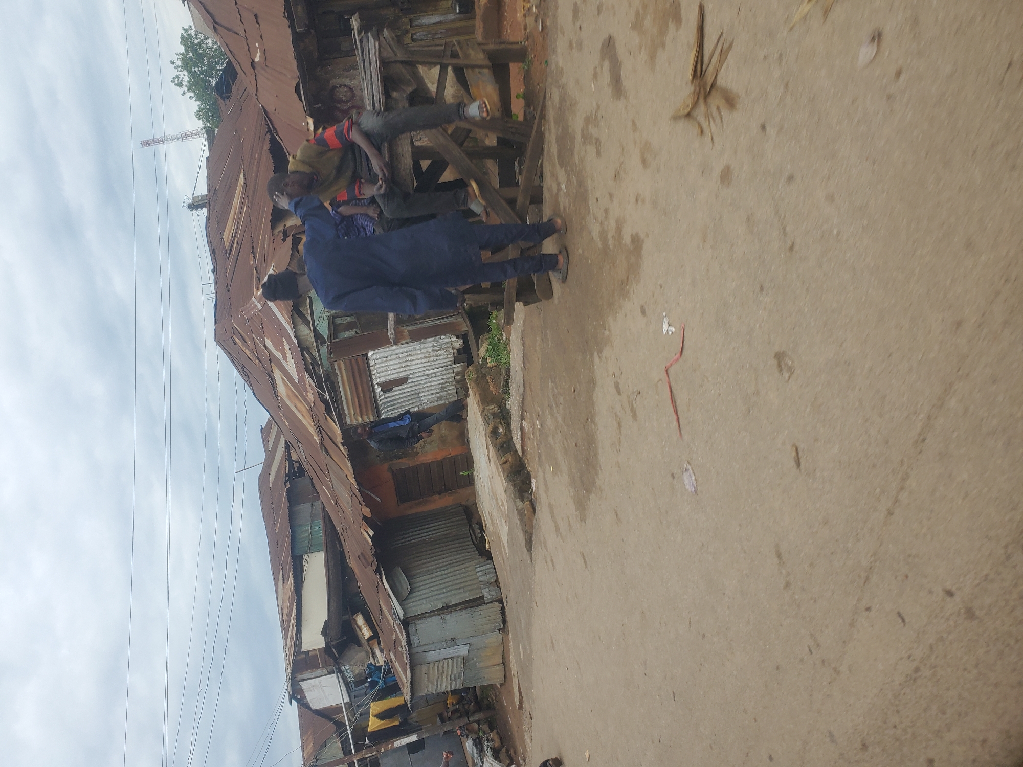 This is a medium shot, taken from a low angle, of a street scene in the city of Jos, Nigeria. The image depicts a crowded urban environment with many informal structures constructed from corrugated iron sheets. In the foreground, a dirt road or path is visible.
In the mid-ground, there are three individuals. Two men are seated on a wooden structure. One man, wearing a yellow and red striped shirt and dark pants, is looking down. The other man, partially obscured, appears to be wearing a blue garment. A third man, dressed in a full blue outfit, stands facing the seated men and appears to be interacting with them, possibly touching the head of the man in the yellow and red shirt.
Behind the people, a collection of ramshackle buildings made of corrugated metal is prominent. These structures appear to be tightly packed together, characteristic of informal settlements. Some laundry is hanging outside one of the structures. The sky above is overcast with grey clouds. The overall impression is one of a densely populated and somewhat impoverished urban area.