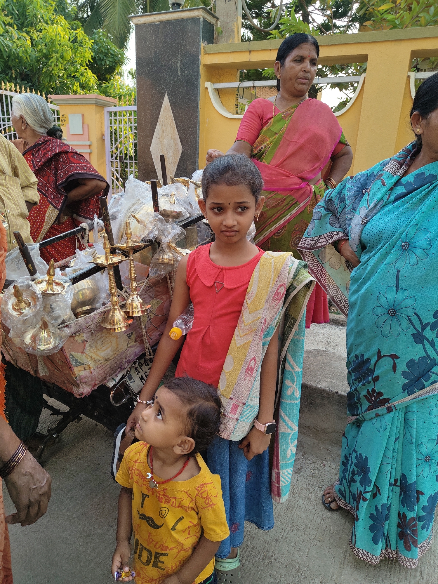 The image, taken in Kammavaripalli, India, shows a group of people on a paved outdoor surface near a building. A young girl in a red shirt and blue skirt stands in the foreground, with a patterned cloth draped over her shoulder. A small child in a yellow shirt with a mustache print stands next to her, looking upwards. Behind them is a cart laden with brass oil lamps, many still wrapped in plastic. To the right of the girl, two women are visible, wearing traditional saris. The first one is wearing a pink and green sari and the second one is wearing a blue sari with floral patterns. To the left of the cart, an older woman in a red and black patterned sari stands near a yellow and white gate. The background includes a building with a gray and yellow facade and trees. The day's lighting is bright and casts shadows, suggesting it's daytime.