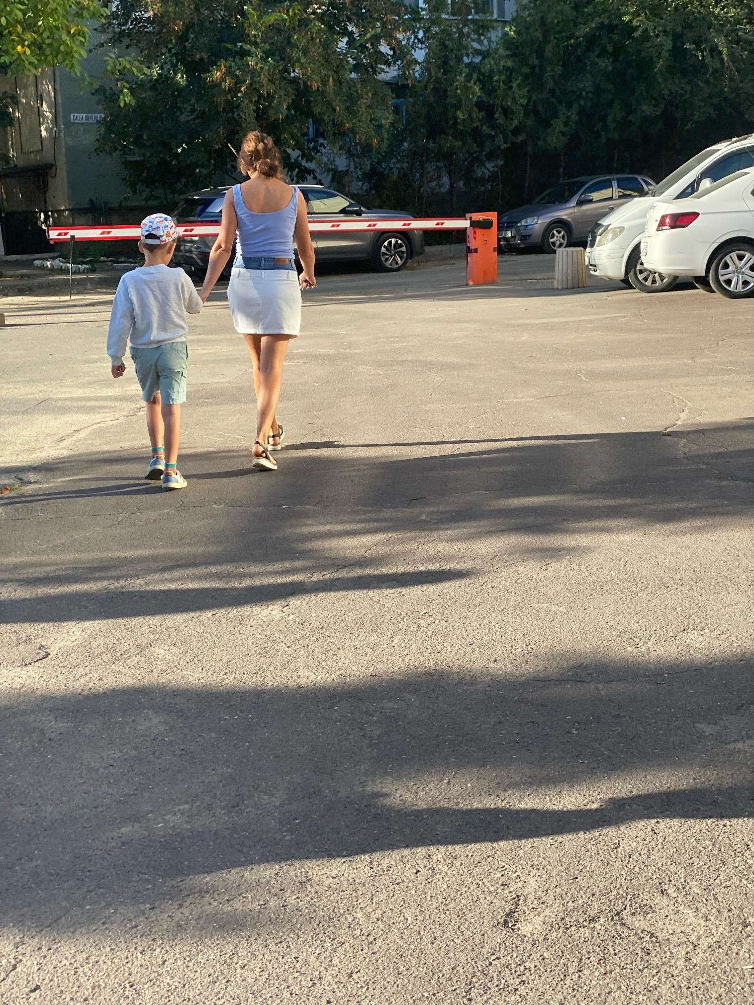 A woman and a young boy, holding hands, walk away from the viewer on a paved street. The woman wears a light blue tank top and a white skirt, and the boy wears a grey sweatshirt and light blue shorts. They are walking towards a red and white barrier gate. In the background, several cars are parked. The scene is illuminated by sunlight, casting long shadows across the street. A street sign visible on a building in the background reads "Calea Unirii".