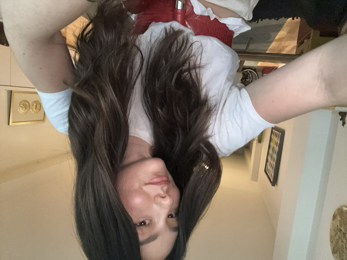A female individual is depicted in a medium close-up, self-portrait photograph within an indoor residential environment. She has long, dark brown, wavy hair, fair skin, and is wearing a white short-sleeved t-shirt with a prominent, wide, textured red belt cinched at her waist. Her right arm is extended, visible on the left of the frame, holding the camera. She maintains direct eye contact with the camera, exhibiting a slight smile. The background, partially out of focus, shows interior decor. To the left, a light-colored wall features a circular gold-toned art piece and a smaller, dark, possibly decorative figurine. Further back, a framed artwork with vertical colorful elements hangs on a wall. To the right, a white furniture unit displays a framed piece of art containing two gold-colored circular objects. Warm ambient lighting is present. The specific location within Mexico City, Mexico, cannot be determined from the visual evidence provided.