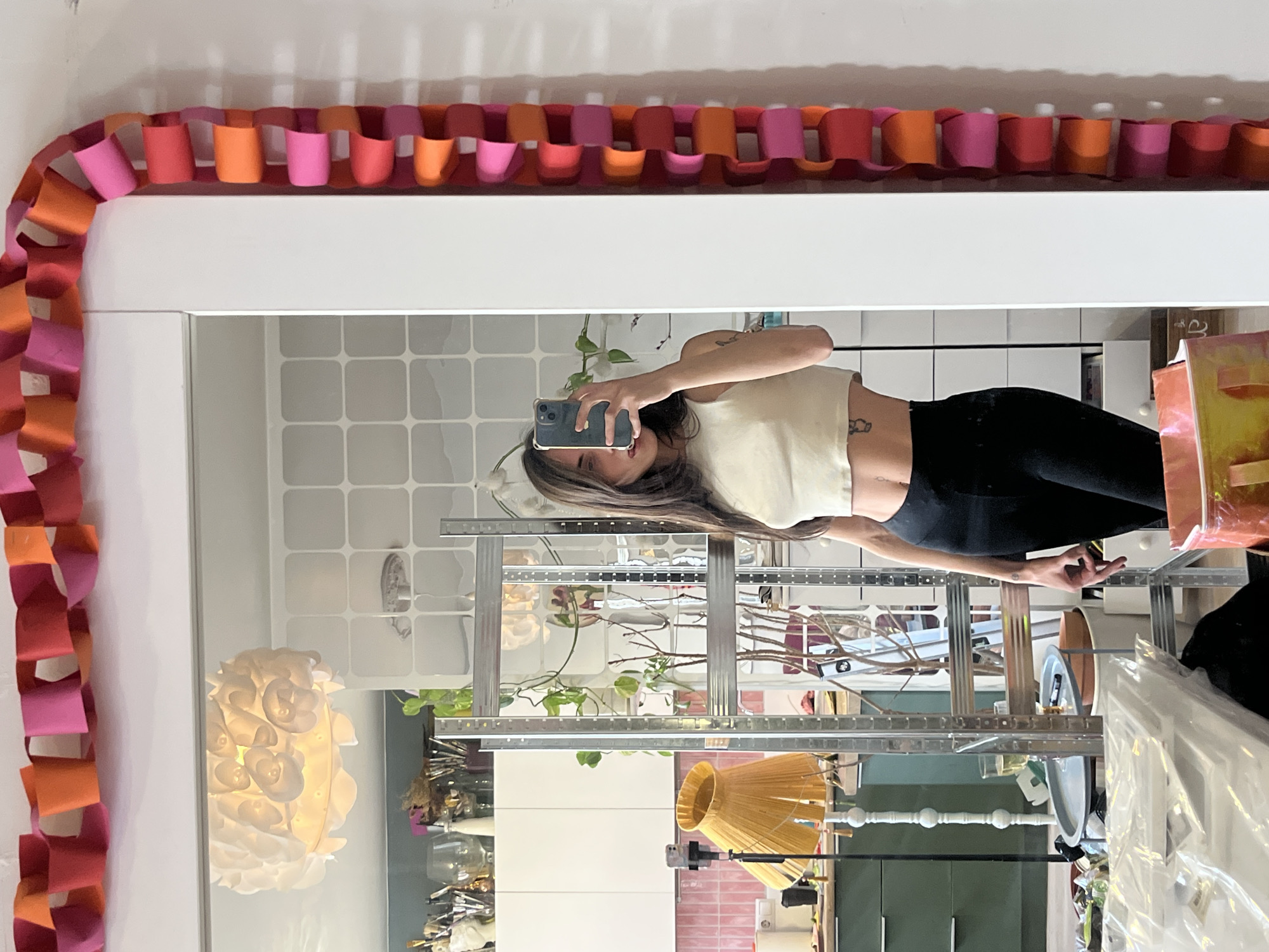 The image depicts a reflection in a mirror decorated with a paper chain of orange and pink. The reflection shows a person holding a smartphone, presumably taking the picture. The person is wearing a light-colored top and black pants. Behind the person in the reflection is a metal shelving unit with various objects, including plants, lamps, and other household items. The background appears to be a room with various surfaces, including tile and painted walls. There is no indication that the photo was taken in Krasnoyarsk, Russia, or that it relates to the city in any way.