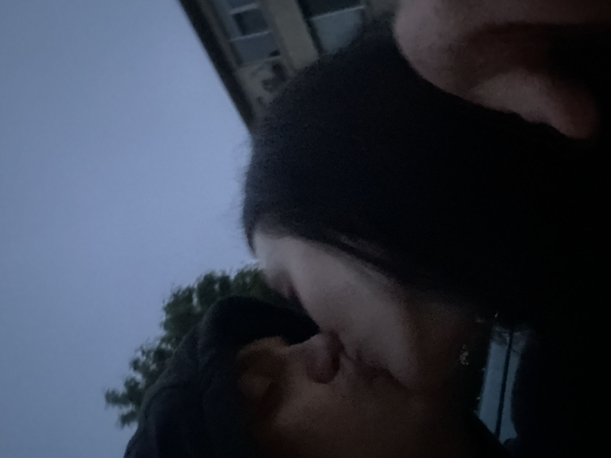Two individuals are intimately kissing, with their faces pressed together. One person is wearing a dark hooded garment. The scene appears to be outdoors, with a building and trees visible in the background. The sky is overcast. There is no confirmed location in Burgas, Bulgaria.