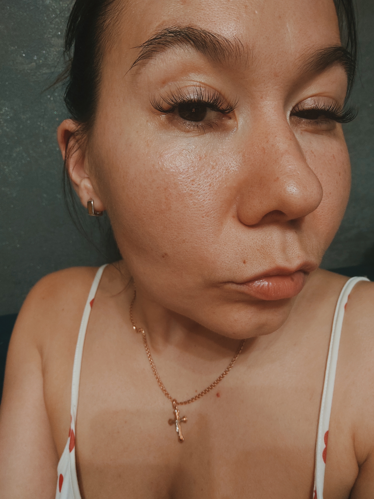 A close-up, medium shot captures the upper torso and face of a young woman. She has her hair pulled back, revealing a small, square, gold hoop earring in her left ear. Her eyes are dark and heavily made up with false eyelashes, and she squints slightly, looking to her right. Her lips are slightly pursed and shiny, with visible lip gloss. A thin, gold chain necklace with a small cross pendant rests on her collarbone. She is wearing a white tank top with a pattern of small red hearts. The background is a textured, dark gray wall. The lighting is warm and appears to be indoor lighting, possibly artificial. The woman's skin has visible pores and some freckles. There is no visible text in the image. The overall impression is one of a casual, personal photograph.