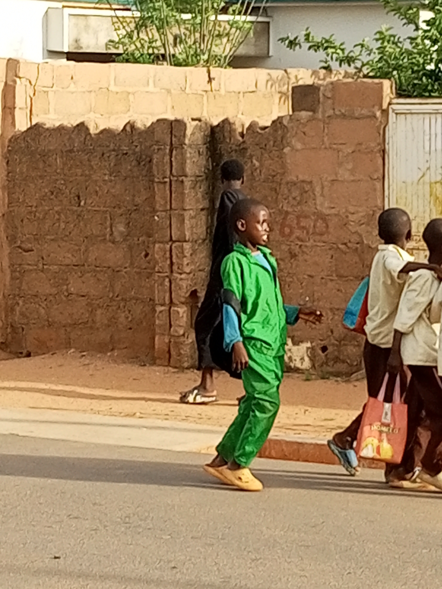 In Tumfure, Nigeria, a group of children is captured walking on a paved road. A young boy, prominently in the foreground, is dressed in a bright green jumpsuit and light brown sandals. He is walking with his head turned and his mouth open, as if speaking or shouting. Behind him, two other children, partially obscured, are also walking. One appears to be wearing a light-colored shirt and dark pants, carrying a blue and red backpack. Another child, dressed in a dark garment, walks slightly ahead and to the left of the boy in green. The background consists of a brick wall, with some vegetation visible above it. The scene is bathed in natural light, suggesting it is daytime.
