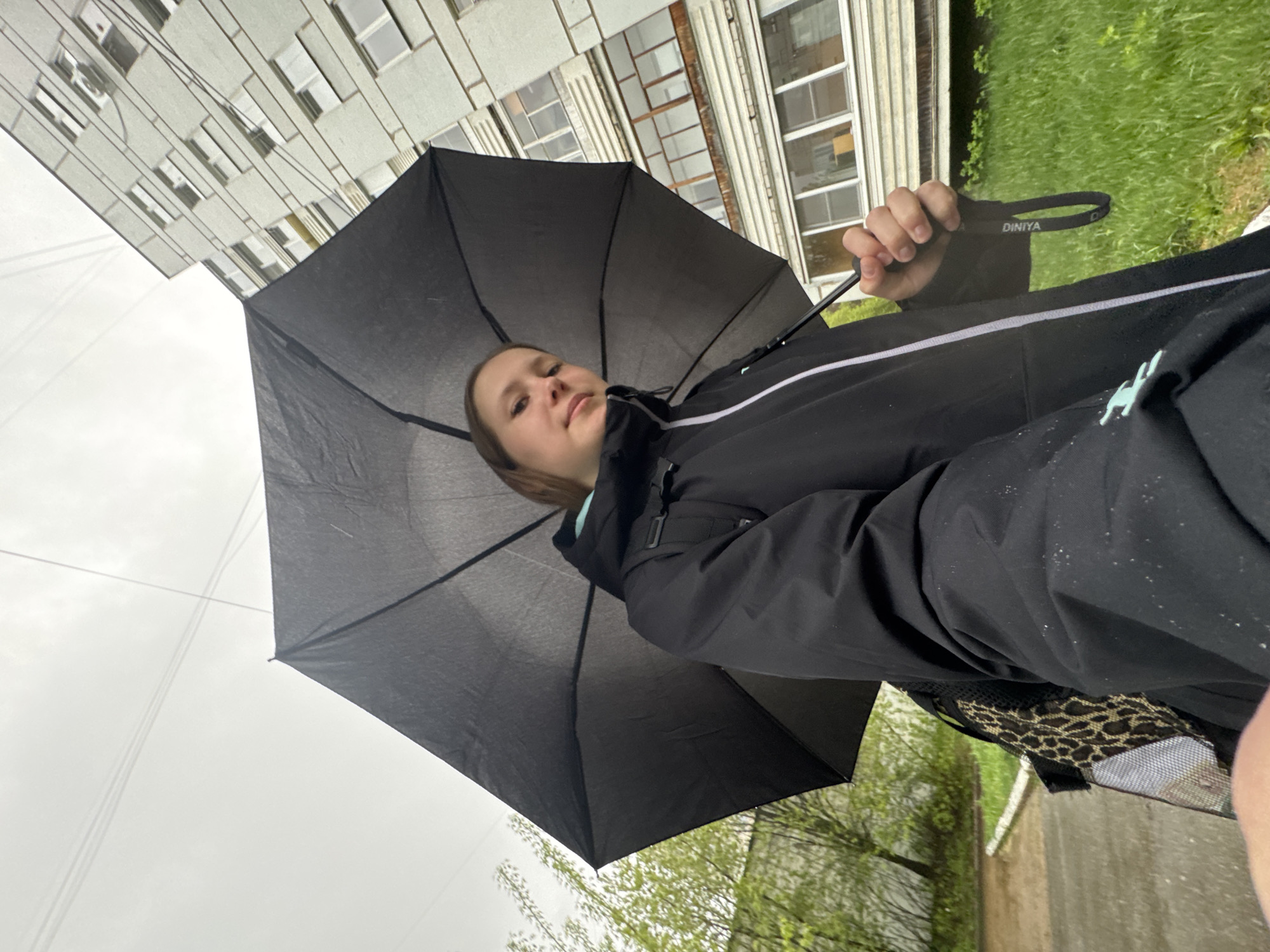 A person stands under a black Dinaya umbrella on a cloudy day in what appears to be the city of Chaikovsky, Russia. The person is facing the camera. A multi-story building is visible in the background along with greenery and a sidewalk.