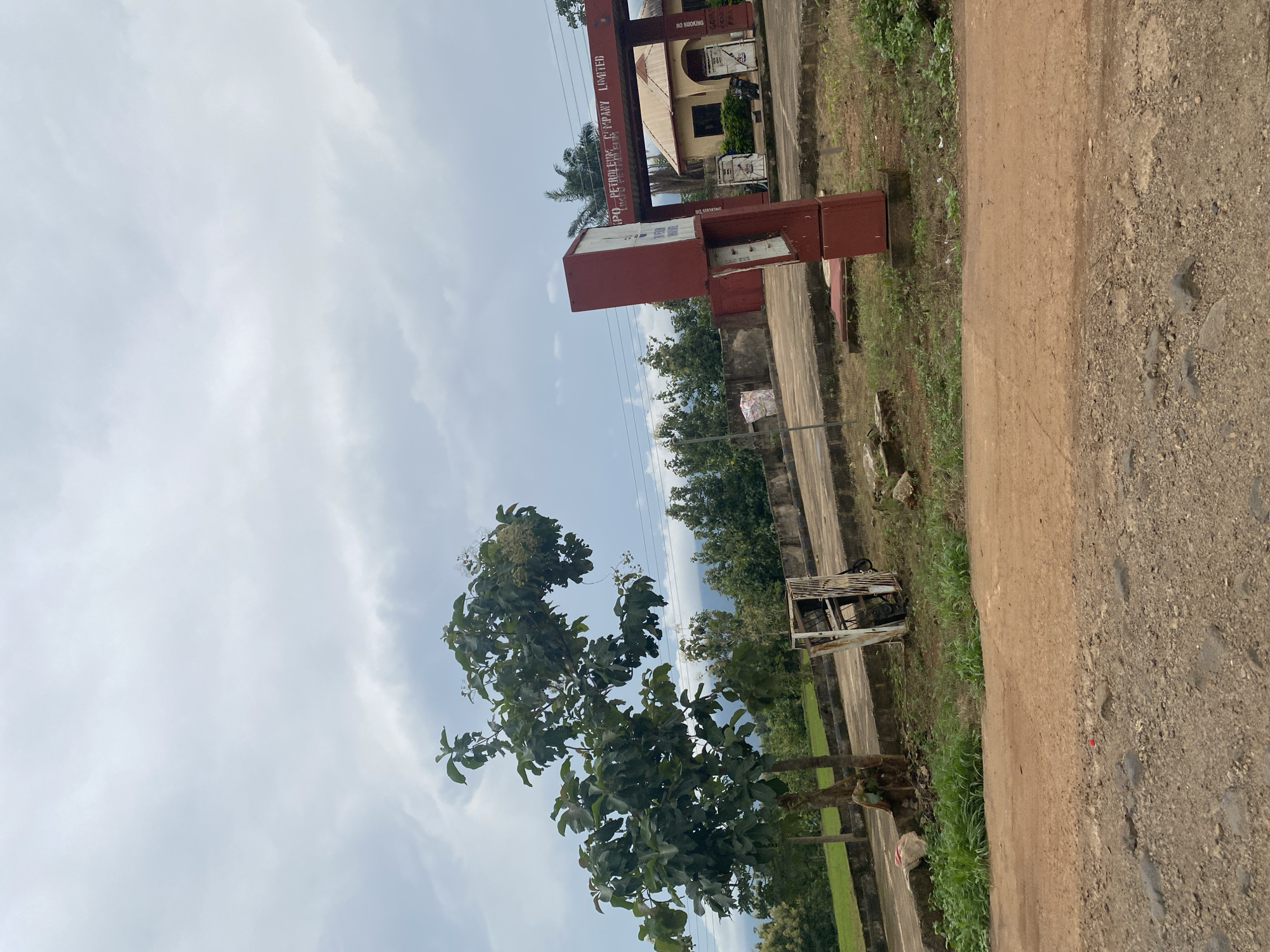 In Karshi, Nigeria, a roadside scene unfolds under a cloudy sky. A large tree with lush green leaves stands to the left, its branches reaching towards the overcast heavens. To the right, a red and white structure, possibly a signpost or a piece of equipment, is situated near a building with a brown roof. Behind these elements, a more distant backdrop of trees and hills suggests a rural or semi-rural setting. The foreground is dominated by a dirt road, with rocks and debris scattered along its surface. A weathered, rusted metal structure, resembling a discarded appliance or piece of machinery, lies on the ground near the tree, adding a touch of disarray to the landscape. The overall impression is one of a slightly neglected but functional area, possibly a gas station or a small business, in a town in Nigeria.