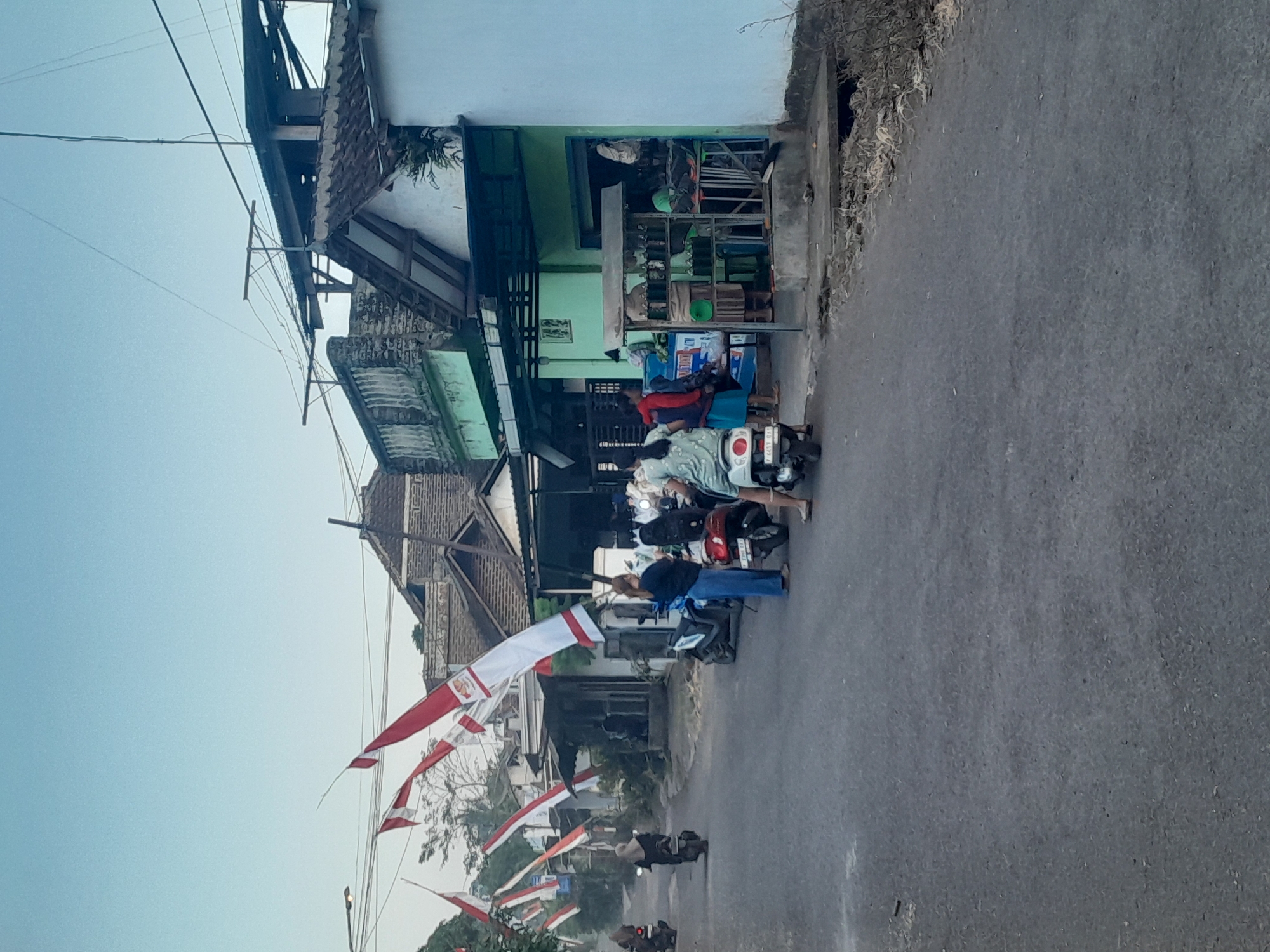 The image depicts a street scene in Sempu, Indonesia, characterized by a paved road lined with buildings and decorated with numerous Indonesian flags. Several individuals are present, some on motorcycles and others standing. In the foreground, two people are visible, one on a white scooter and another standing next to it, wearing a blue top and pants. To the right, a small storefront is open, displaying shelves with numerous bottles, possibly containing fuel or beverages, and a person can be seen inside. The buildings appear to be low-rise, with tiled roofs and a mix of weathered and freshly painted facades, including one painted in a light green. The street is relatively narrow, with electrical wires strung overhead. Indonesian flags, red and white, are prominently displayed, attached to poles and hanging across the street, suggesting a national celebration or event. The overall atmosphere is one of daily life and local commerce.