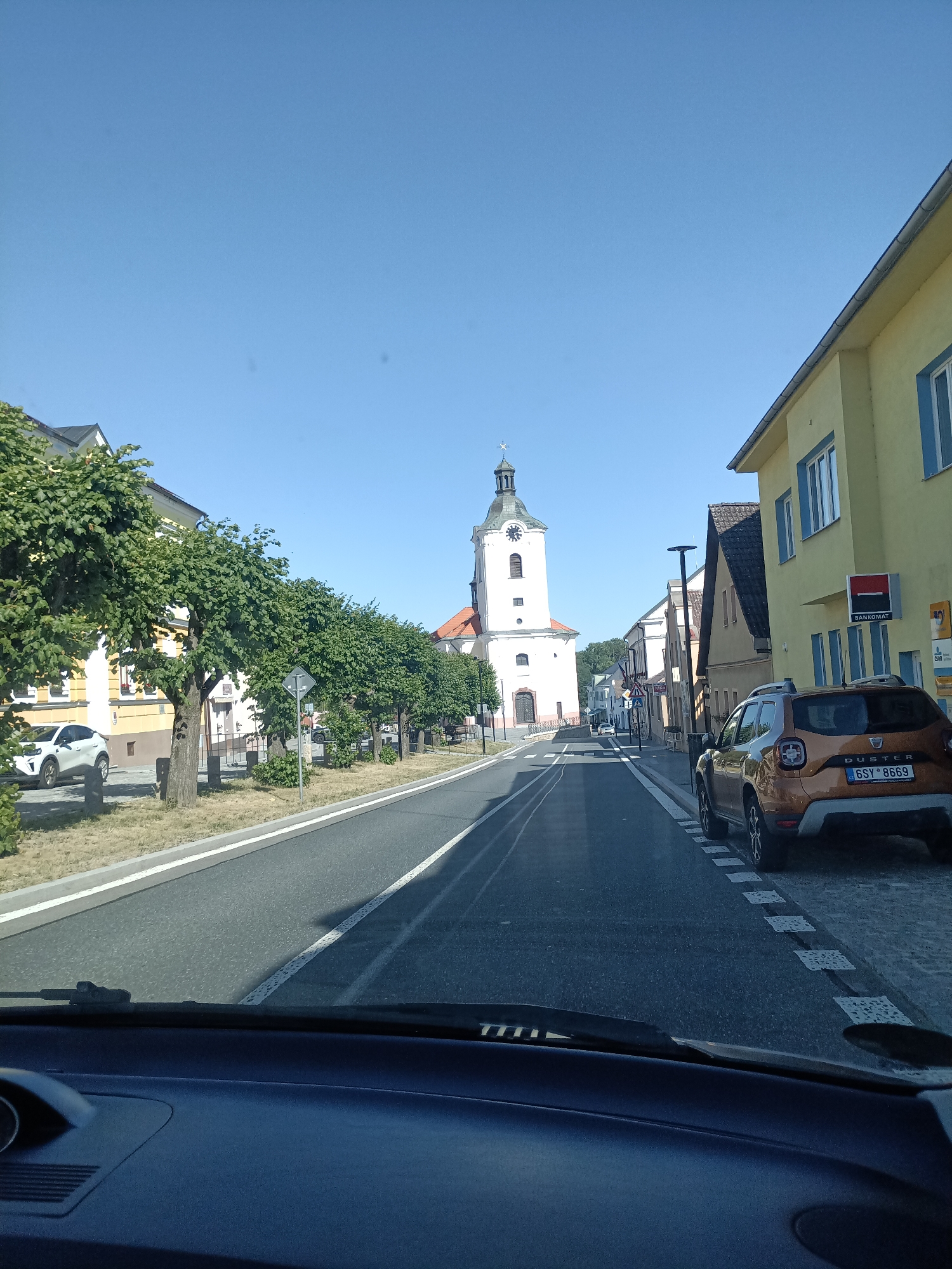 The image shows a street scene in Dalovy, Czechia, on a clear, sunny day. In the foreground, the dashboard and windshield of a car are visible. The street is paved, with a white line dividing the lanes. On the right side of the street, there is a yellow building with blue-trimmed windows and a sign reading "BANKOMAT". A brown Dacia Duster is parked on the right side of the road.
Further down the street, a white church tower with a clock is prominent. The church has a red roof and is surrounded by trees. Several other buildings line the street, and a row of green trees with dense foliage stands along the left side of the road. A white car is parked on the left side. The sky is a clear, bright blue. The overall impression is a quiet, well-maintained street in a European town.