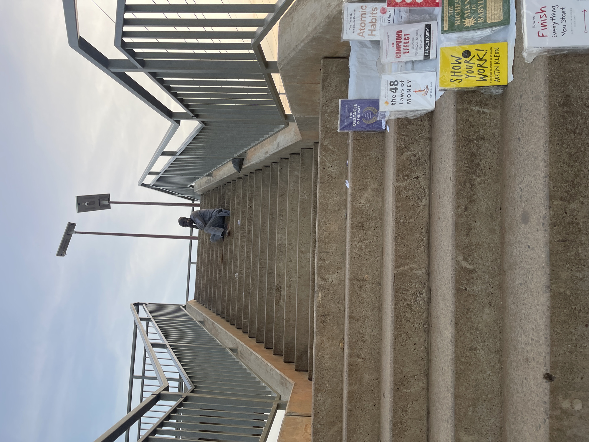 A low-angle shot depicts a staircase with a metal railing, ascending to an overpass with streetlights, seemingly in Galadimawa Village, Nigeria. A person sits on the steps midway up. A selection of self-help books is displayed on the steps to the right, including "Atomic Habits," "The Compound Effect," "The 48 Laws of Money," "Show Your Work!" and "The Richest Man in Babylon." A book titled "Finish" is also visible. The books are possibly for sale. The sky is cloudy.