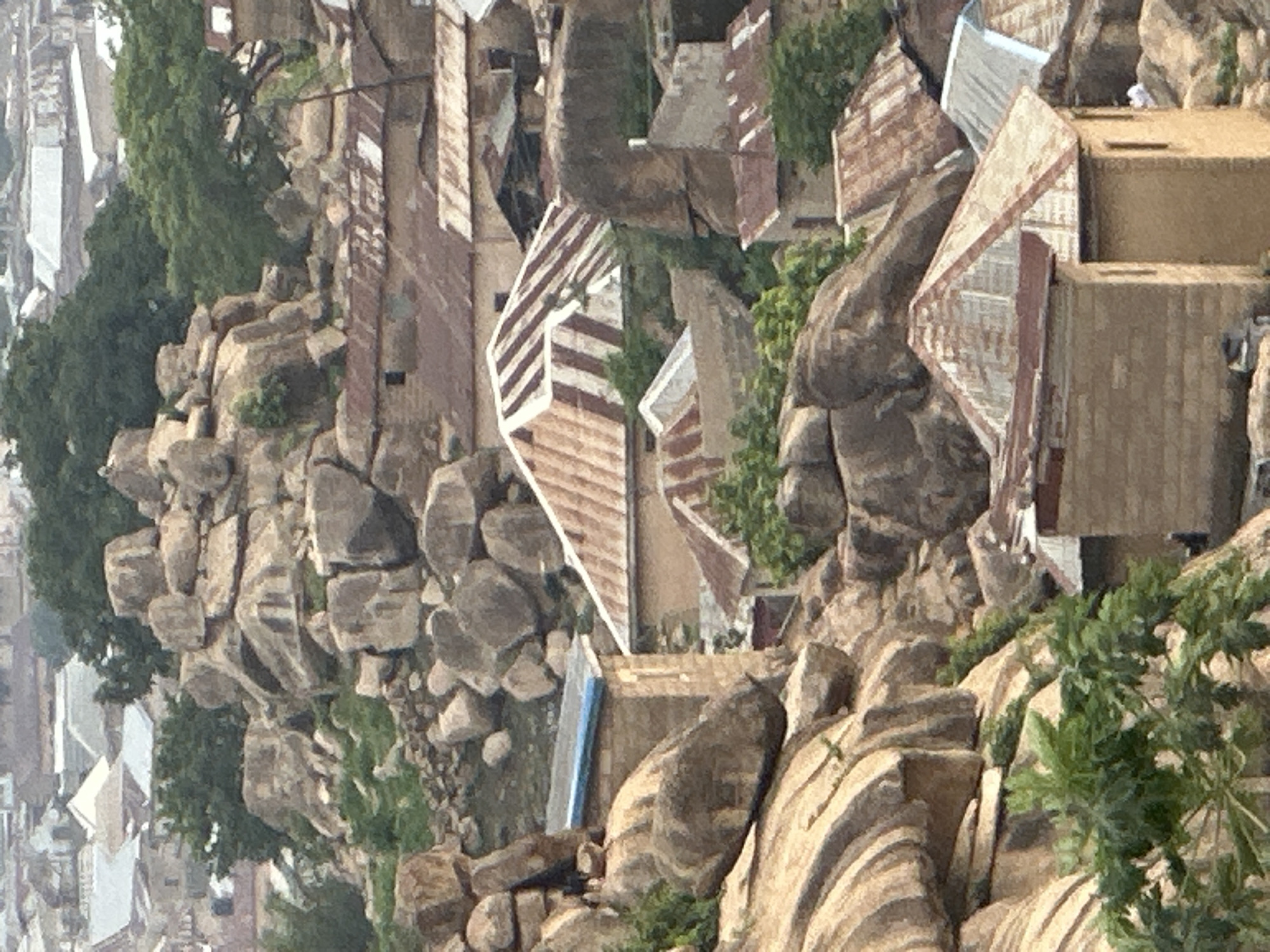 The image displays an elevated view of a densely populated area in Jos, Nigeria, characterized by human settlements built among and around prominent, large rock formations. Numerous structures, predominantly houses, feature various types of roofs, including corrugated iron sheets in shades of brown, red, and silver. Some roofs appear aged or rusty, while others are newer and metallic. The buildings themselves exhibit earthy-toned walls, likely constructed from mud-brick or concrete, and are positioned in close proximity, often directly abutting or nestled between the natural rock outcrops.
The scene is dominated by massive, smooth, rounded granite boulders that rise significantly above the surrounding terrain, with human habitation intricately adapting to this challenging landscape. Scattered green trees and lush vegetation are interspersed among the rocks and buildings, providing pockets of natural greenery. The overall impression is one of a community living in close harmony and adaptation to a rugged, rocky environment.
The time of day appears to be daytime, with soft, diffused lighting suggesting an overcast sky or hazy conditions. There are no people visible, and no specific activities or events can be discerned. No visible text is present in the image.