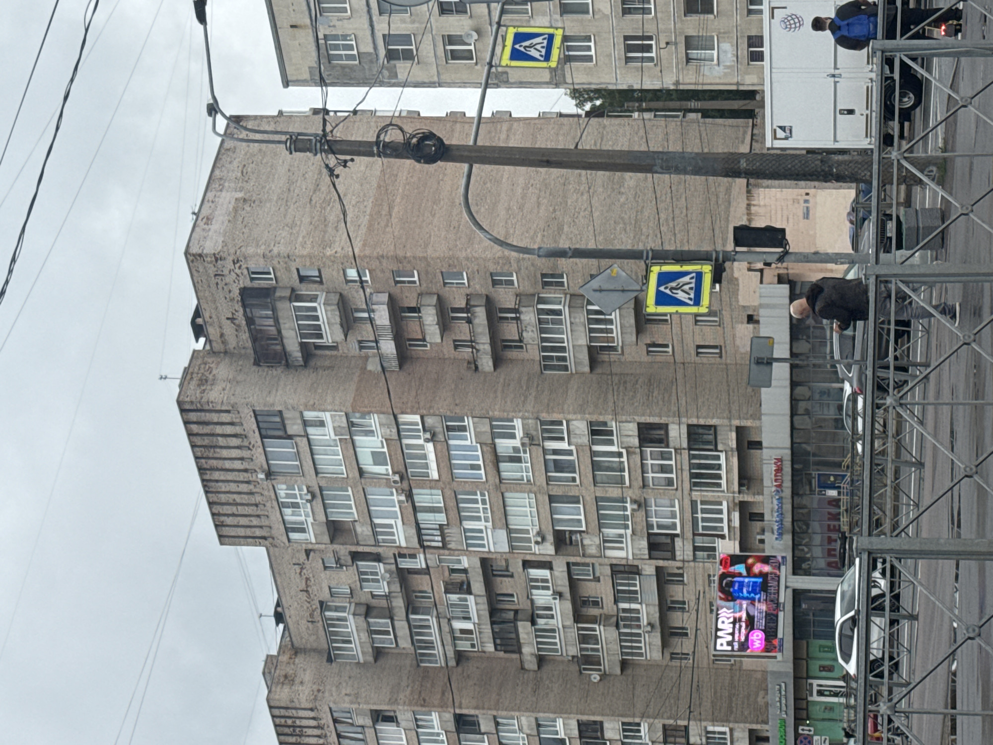 This media file shows an urban scene in Saint Petersburg, Russia, with a focus on a large, weathered brick apartment building. The building features numerous windows, many with balconies or enclosed balconies, and is showing signs of age and urban grime. Overhead power lines crisscross the frame, adding to the utilitarian feel of the environment.
In the foreground, a metal scaffolding structure is visible, suggesting ongoing construction or maintenance work. A pedestrian crossing sign, recognizable by its blue and yellow diamond shape with a white pedestrian symbol, is mounted on a pole. Another smaller, grey diamond-shaped sign is also present.
Several people are visible in the scene. One person, wearing a blue backpack, stands near a white truck, possibly a delivery vehicle. Another individual, a man with a shaved head, is seen near a parked car. The presence of these individuals suggests the area is a public space with daily activity.
A bright, colorful advertisement billboard is prominently displayed on the side of a building in the middle ground, featuring bold text and imagery, which stands in contrast to the muted tones of the architecture. Some of the visible text on the billboard includes "PWR" and "wb". Below the billboard, there appear to be shops or commercial establishments with signage.
The sky is overcast, indicating a cloudy or possibly rainy day, and the overall lighting suggests it is daytime. The perspective is tilted, as if the camera was held at an angle. The scene captures a slice of everyday life in a Russian city, marked by its architecture, infrastructure, and the presence of people going about their business.