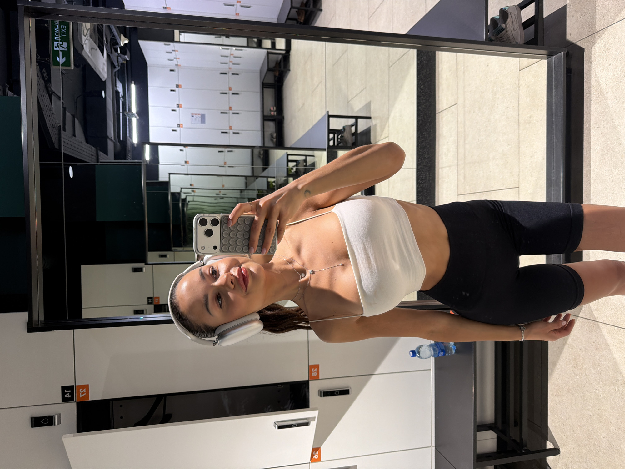 The image depicts a young adult female taking a mirror selfie in a modern gym locker room, located in the city of Moscow, Russia. The woman, with light skin and dark hair pulled back, is attired in a white spaghetti-strap crop top, black cycling shorts, and white over-ear headphones (Apple AirPods Max). She holds a smartphone, encased in a grey "pop-it" style cover, in her right hand, and is smiling at her reflection. Her visible accessories include a two-layered silver necklace with circular pendants, a silver bracelet on her left wrist, and a small crescent moon tattoo on her right forearm.
To her immediate left, a portion of a white locker with a dark handle and an orange number "8c" (reversed) is visible. A dark gray bench is positioned below this locker, holding a blue-capped water bottle. The large mirror in which the selfie is taken reflects the clean, well-lit locker room, showing multiple rows of white lockers with dark handles and orange numbering, along with dark gray benches and a light-tiled floor. The ceiling, visible in the reflection, features exposed ducts and fluorescent lighting. A green exit sign, displaying both Cyrillic "ВЫХОД" (VYKHOD) and English "EXIT," is clearly visible on the far wall in the mirror's reflection, indicating a Russian-speaking environment.