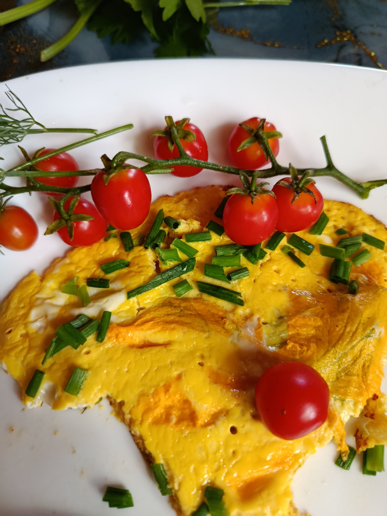 A close-up, overhead shot captures a white plate holding a partially eaten omelet. The omelet is bright yellow with visible white egg whites and scattered chopped green chives. Several cherry tomatoes on the vine are artfully arranged above the omelet, with one single cherry tomato resting on the edge of the omelet. A sprig of dill is also present. The background features dark green leafy vegetables and a dark, textured surface, suggesting an outdoor or rustic setting, possibly in the city of Vereya, Russia.