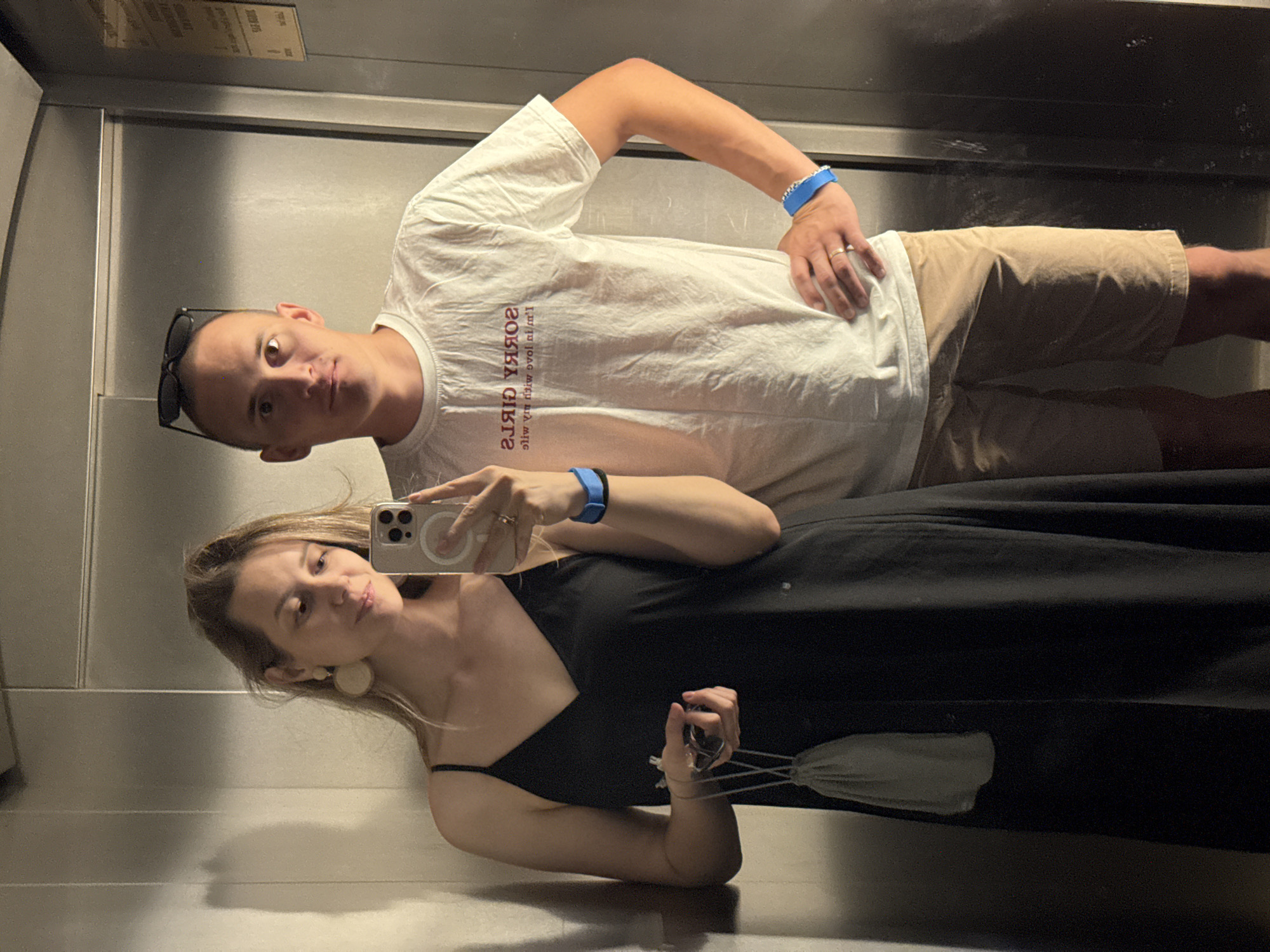 This full-shot image captures two individuals, one female and one male, standing in what appears to be an elevator or a room with reflective metallic walls, as contextually located in Nebug, Russia. Both subjects are looking towards the camera, suggesting a mirror selfie.
The female, positioned on the left, has light brown hair and is wearing a dark, possibly black, V-neck camisole dress. She has large, light-colored circular earrings and a gold-colored ring on the ring finger of her right hand. Her left wrist bears a blue wristband with a darker blue segment. She holds a light-colored smartphone with a visible multi-lens camera array and a circular MagSafe ring on its back, pointed towards the mirror. In her left hand, she holds a small, light grey drawstring pouch with a dark object inside. Her facial expression is a slight smile, and her gaze is directed towards the phone screen.
The male, positioned on the right, has short hair and sunglasses resting on his head. He is wearing a white crew-neck t-shirt with mirrored red text on the chest, which, when corrected, reads: "SORRY GIRLS I'M IN LOVE WITH MY WIFE". He wears khaki shorts and a blue wristband on his right wrist, similar to the female's. A gold-colored ring is visible on the ring finger of his left hand. His right hand rests on his hip, and his gaze is directed straight forward into the mirror.
The environment consists of light grey, metallic-paneled walls with vertical seams, indicative of elevator doors or paneling. On the far right wall, a faded and yellowed rectangular sign with unreadable text is affixed. The lighting casts subtle shadows, particularly on the left wall behind the female.