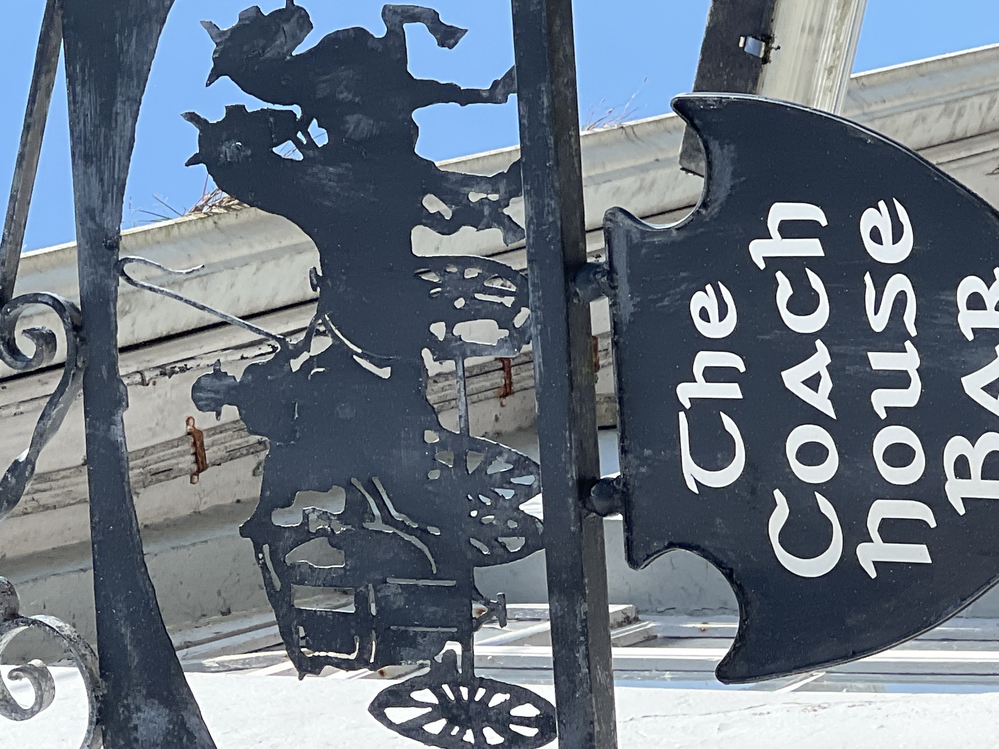 The image shows a black metal sign hanging from an ornate metal bracket, depicting a horse-drawn carriage with two horses. Below the carriage silhouette, a black shield-shaped sign with white lettering reads "The Coach house BAR". The sign appears to be located on a building in a European setting, likely a town or city. The background shows a white building with wooden trim and a clear blue sky. Based on contextual clues from other images, this sign is located on The Coach House Bar in Donegal Town, County Donegal, Ireland.