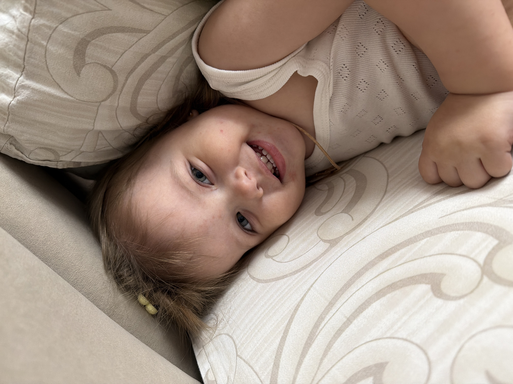 A young girl is lying down on a light-colored patterned cushion, possibly a sofa. She is smiling and looking at the camera. She is wearing a light-colored shirt. Two yellow hair clips are visible in her hair. The location is unknown, but based on the prompt, it may be Reshetnikovo, Russia.