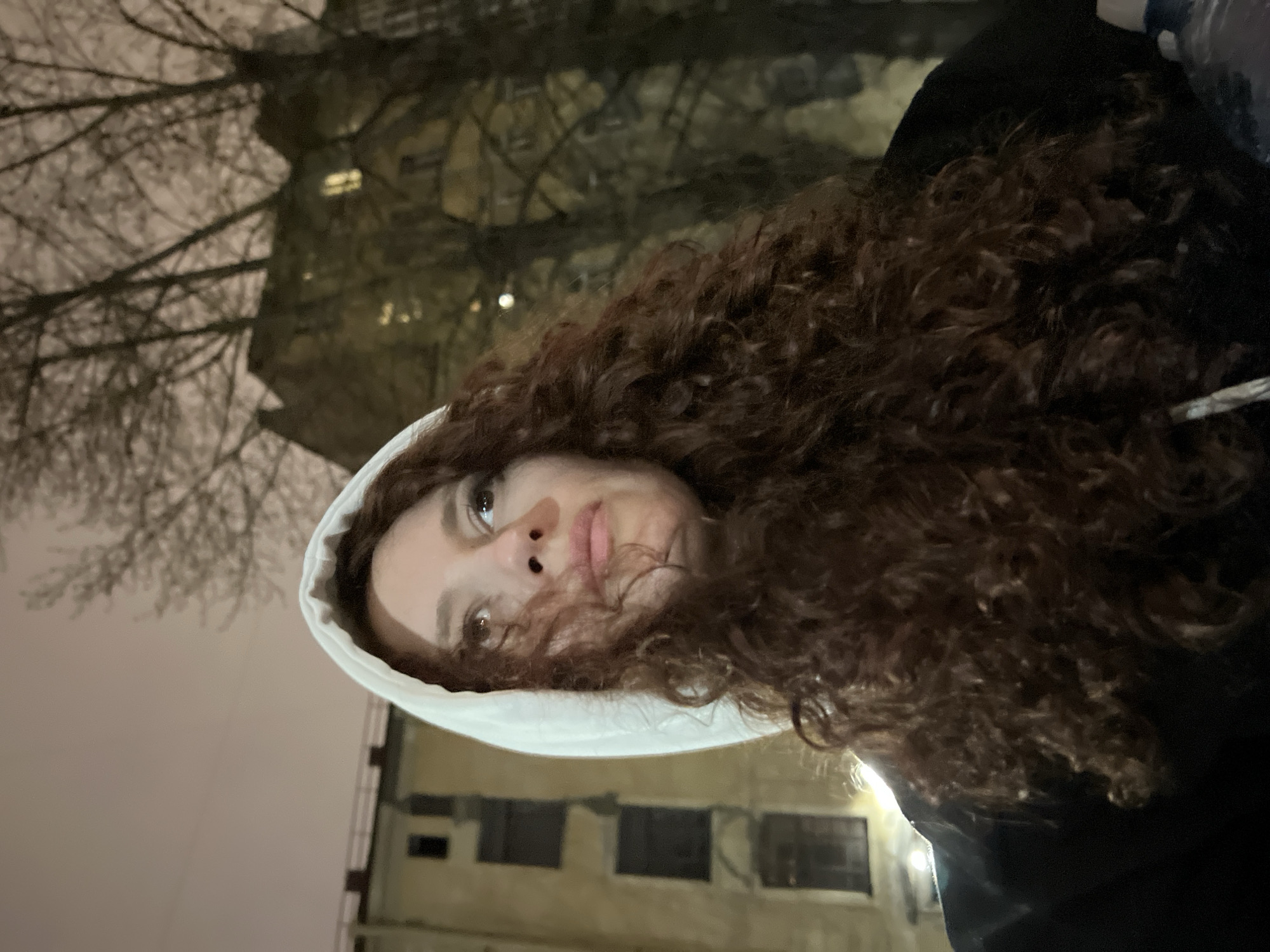A woman with curly brown hair, wearing a white hooded sweatshirt and a dark jacket, is photographed from a low angle at night. She looks upwards with a contemplative expression. The background features a bare tree silhouetted against a pale sky and the facade of a building with windows visible. Based on architectural styles, this location is likely in Saint Petersburg, Russia.