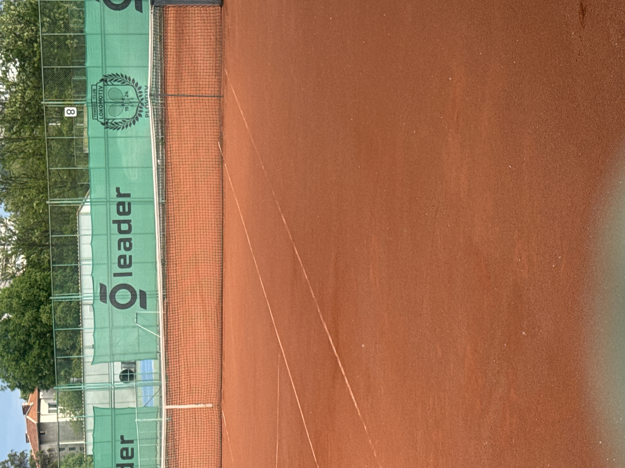The image shows a tennis court in Plovdiv, Bulgaria. The court is clay, with typical white lines marking the boundaries. A net divides the court in half. In the background, a green barrier runs along the back and sides of the court, featuring the word "Leader" in white letters and the logo of "Tennis Club Lokomotiv Plovdiv 1924." The barrier also has the number "8" visible. Beyond the barrier are trees and a building, indicating the court is located in an urban area. The perspective of the photograph is from ground level on the tennis court. The sky is visible through the trees.