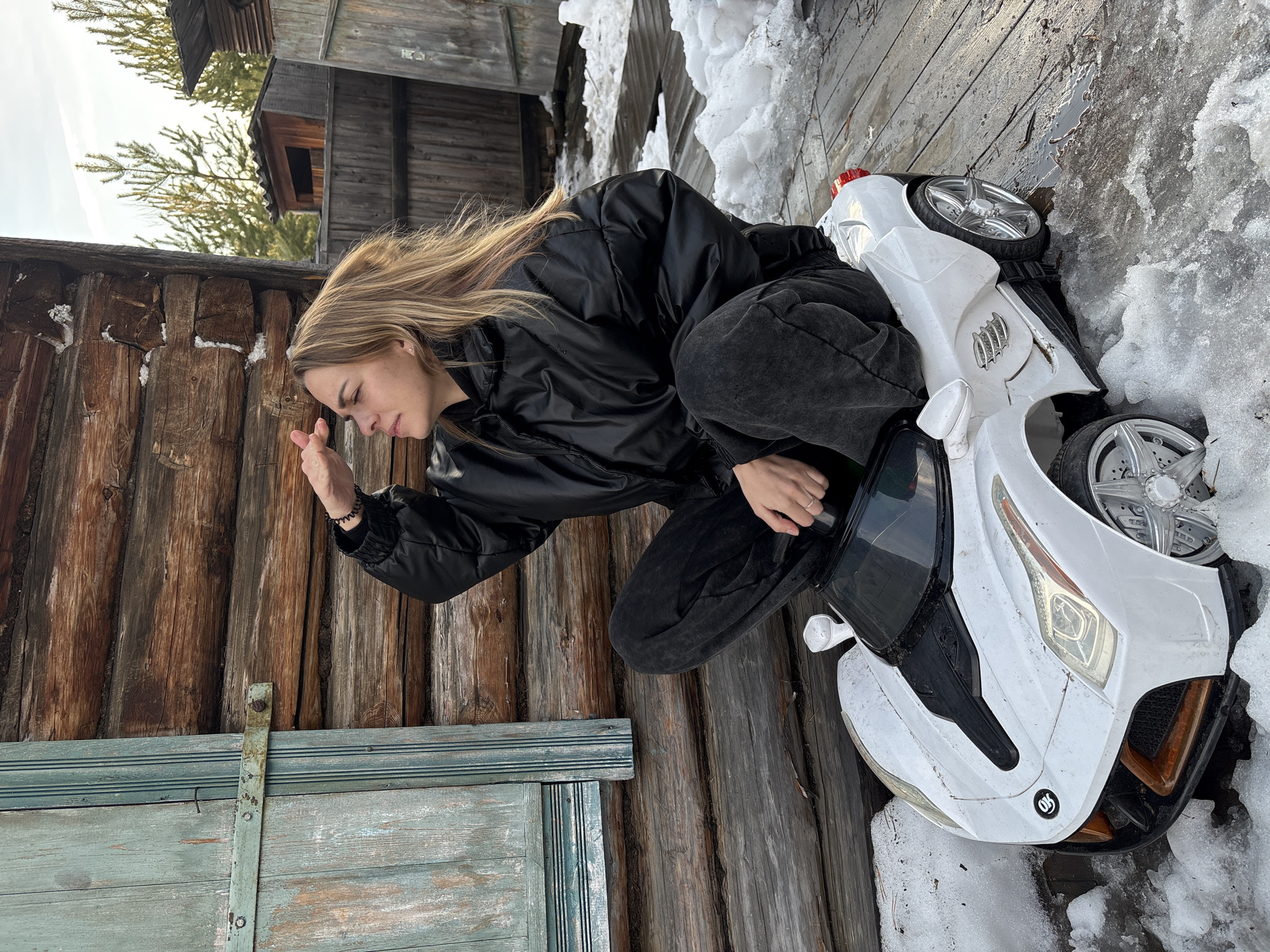 A woman with long blonde hair, wearing a black jacket and dark-colored pants, is crouching next to a white toy car outdoors. The woman is looking towards a wooden structure with a closed, weathered green door. Snow is visible on the ground, and the area appears to be located near wooden buildings. The scene is likely in a rural or semi-rural location, evidenced by the building materials and surrounding environment. The image does not provide enough information to confirm a specific location in Zheleznodorozhny, Russia.