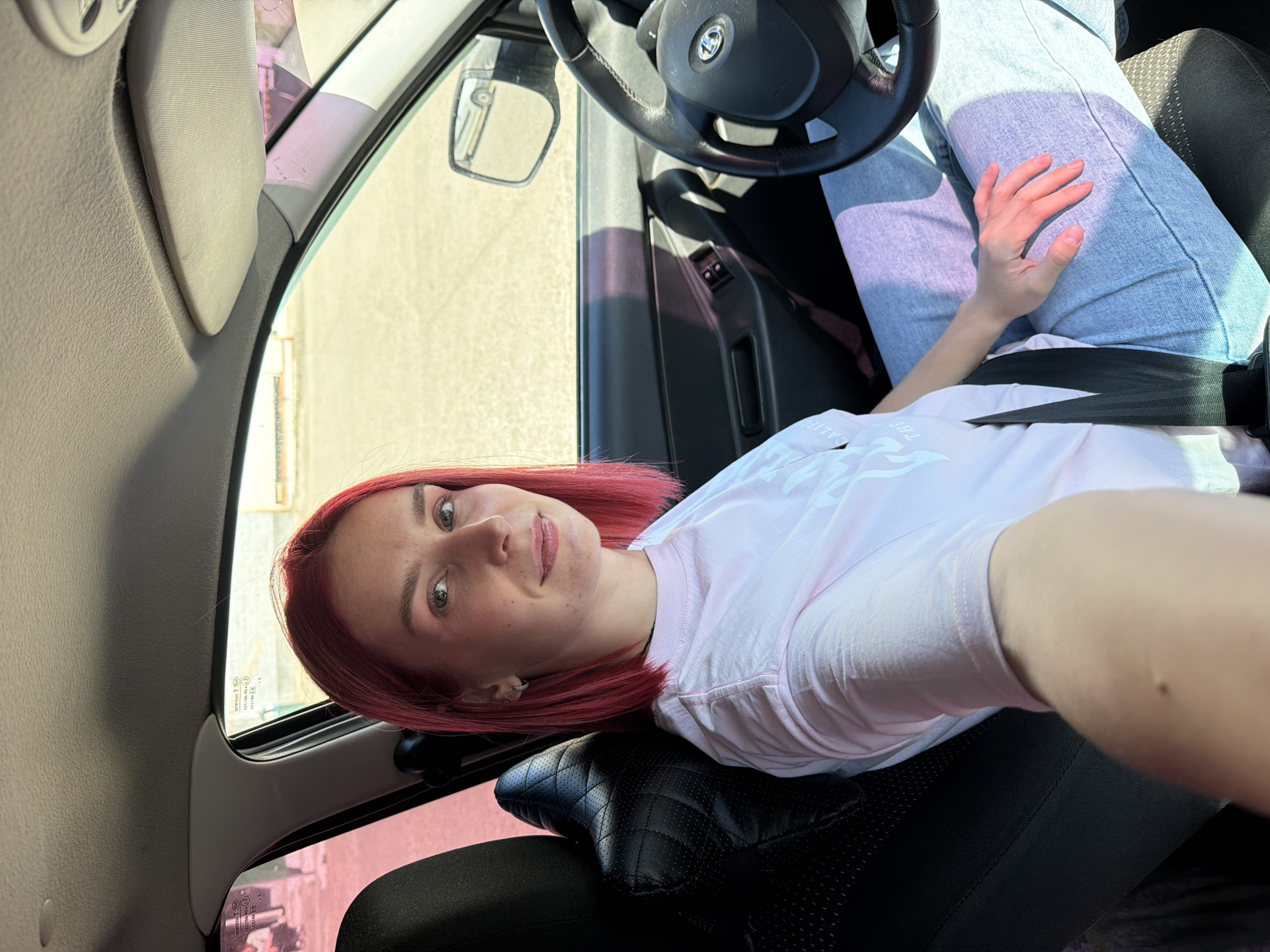 A young woman with short, dyed red hair is seated in the driver's seat of a car. She is wearing a light pink t-shirt and light blue jeans. A seatbelt is visible across her body. The interior of the car is visible, including the steering wheel, dashboard, and door panel. The windows are tinted. The background appears to be an outdoor environment with a paved surface and some buildings. The image was taken in Chaikovsky, Russia.