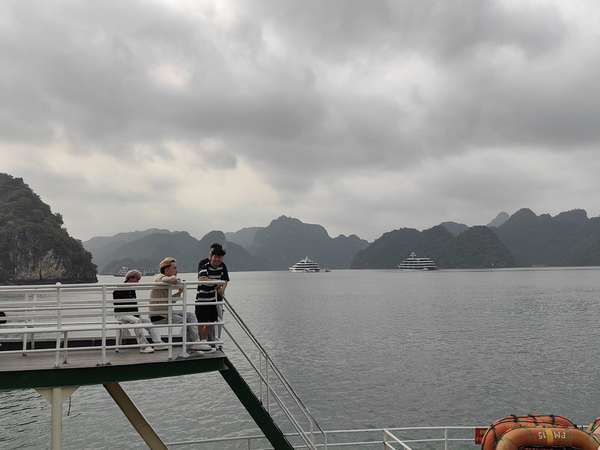 The image shows three people standing on the deck of a boat. Two people are sitting, and one person is standing. They appear to be looking out at the water. In the background, there are islands and two boats on the water. The weather is overcast. The image was taken in Tuan Chau Ward, Vietnam.