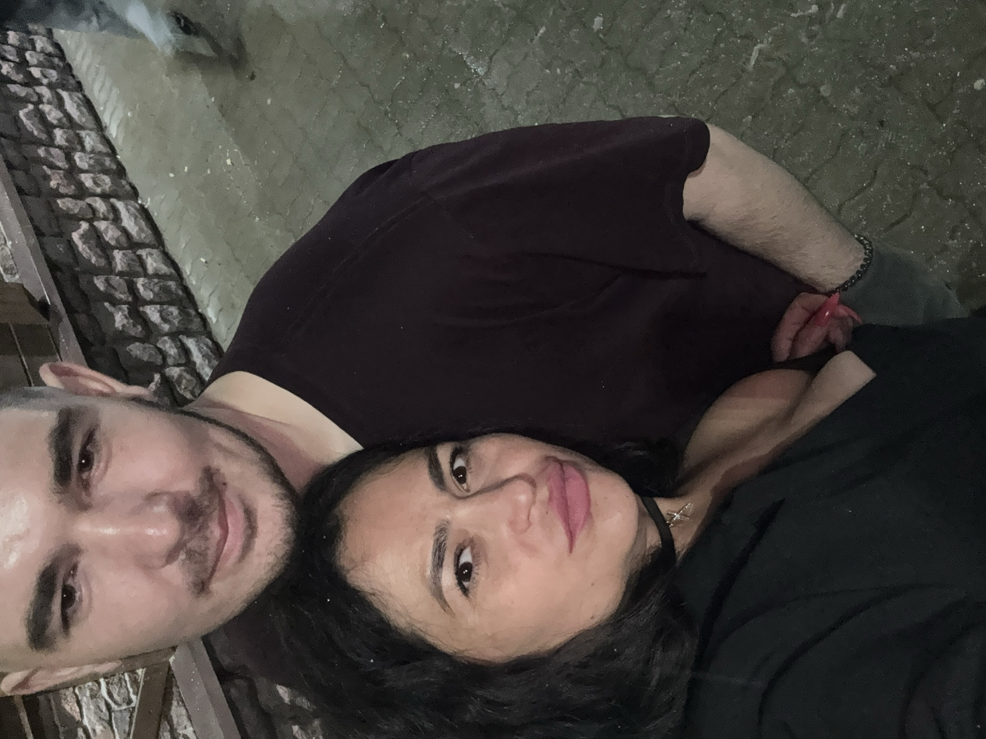 A selfie of a man and a woman in Chaikovsky, Russia. The man has dark hair, a short beard, and a purple shirt. The woman has dark hair, a black top, and a choker necklace. They are standing on a brick-paved street.