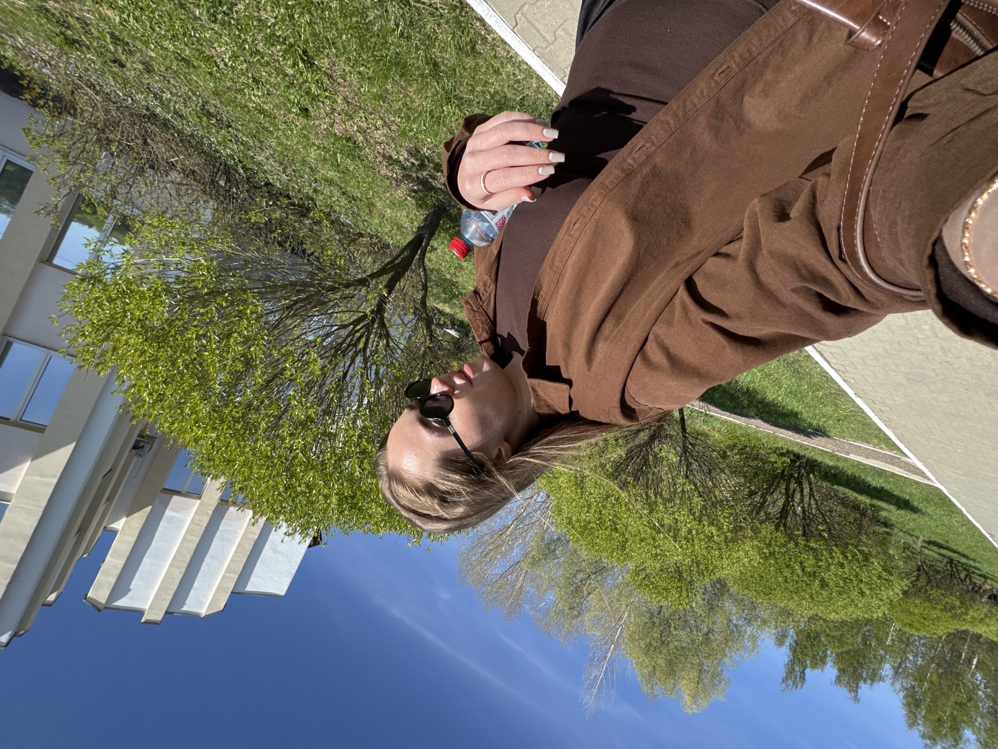 A young woman wearing sunglasses, a brown top, and a brown jacket is sitting outside. She is holding a water bottle in her left hand, on which she wears a gold ring. The sky is blue, and there are trees with green leaves surrounding her. In the background, there is a white building. The location is identified as Chaikovsky, Russia.