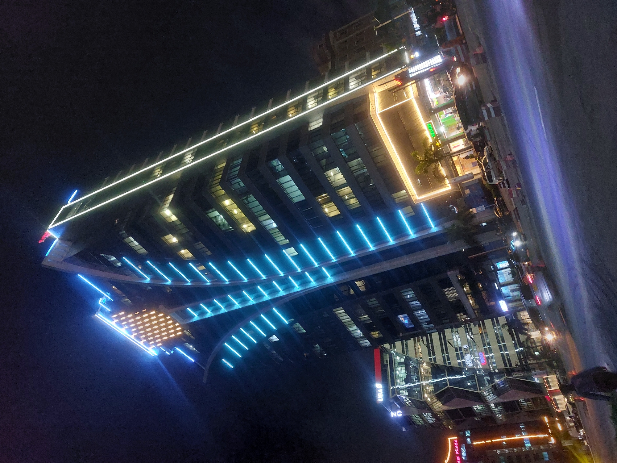 The image is of a building with blue and white lights at night. The building is located on a busy street with people walking by. There are cars parked along the side of the road. The building has a modern design and is lit up with bright lights. The location of the building is in Dar es Salaam, Tanzania.