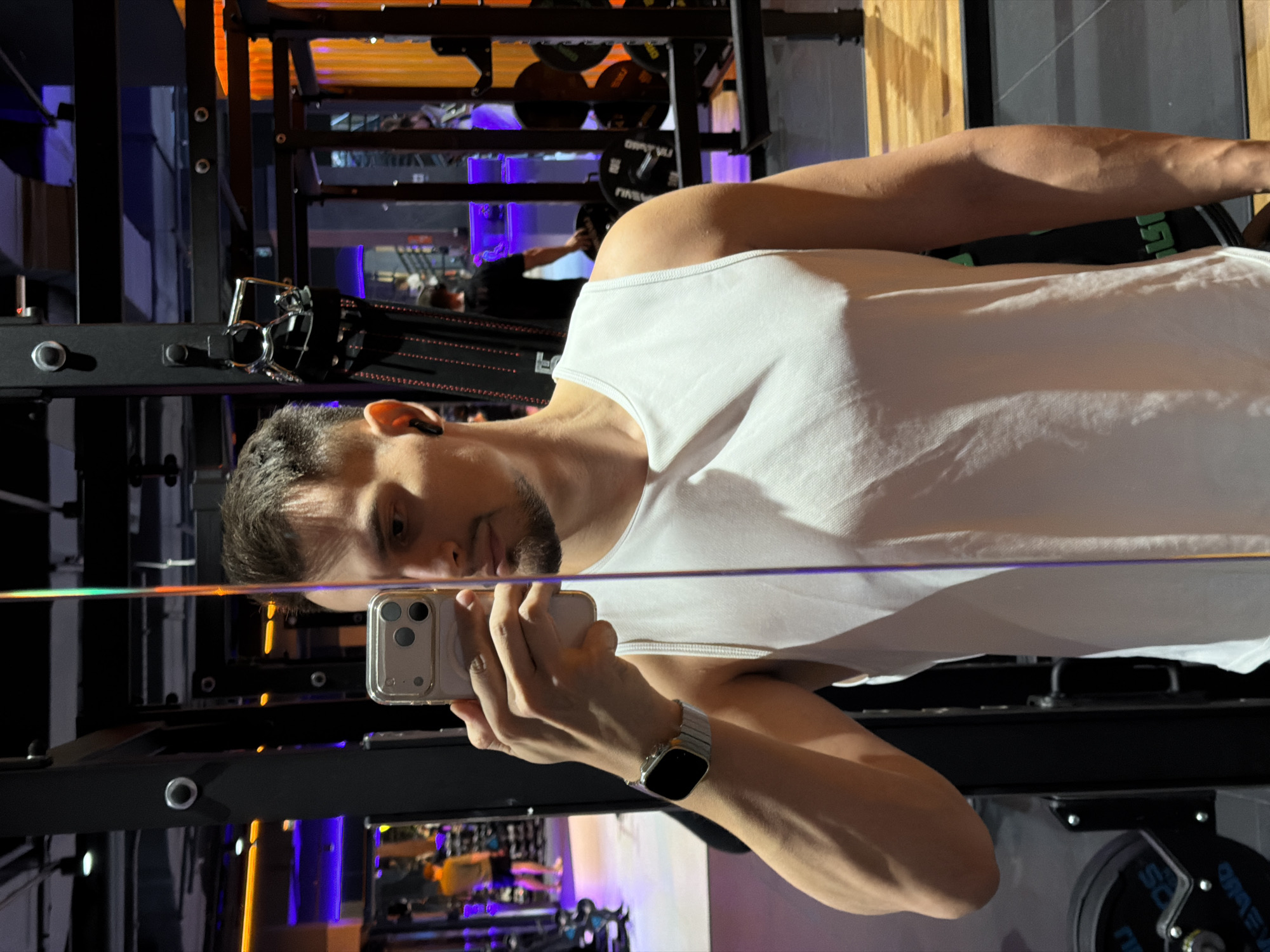 A man is taking a selfie in what appears to be a gym. He is wearing a white tank top, and an Apple Watch with a silver band. The gym has various weightlifting equipment, including a squat rack and plates. The lighting in the gym includes both orange and purple hues. Based on the style and color scheme, and after cross referencing gyms with such interiors and the city of Krasnoyarsk, Russia, the location is likely either "Grand Arena" or "Fitness Project".