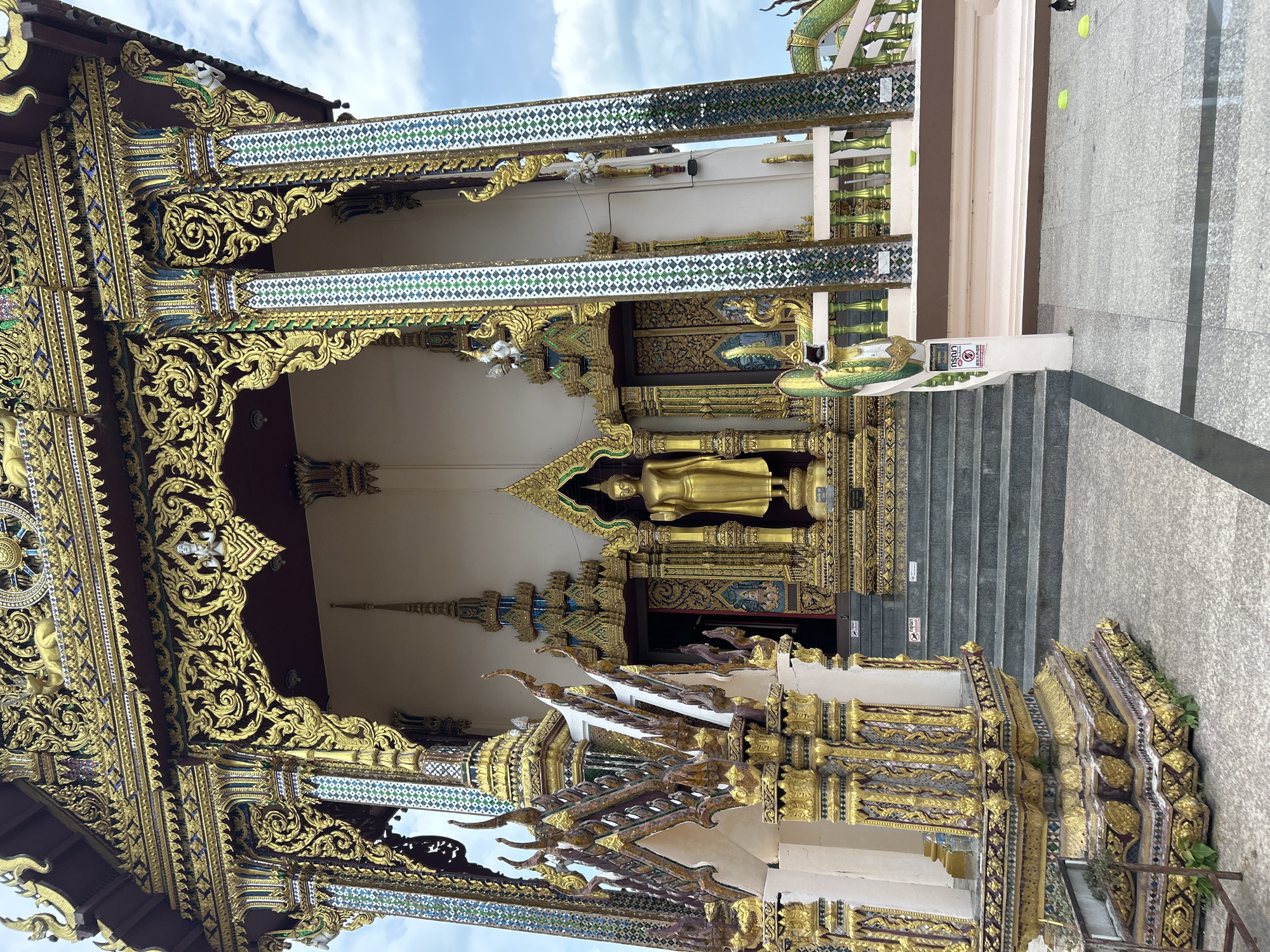 A golden Buddha statue stands prominently on a raised platform in front of a richly decorated temple entrance in Ko Samui District, Thailand. The temple is adorned with intricate gold carvings, colorful mosaic tiles, and ornate architectural elements. A set of grey stone stairs leads up to the temple entrance, with decorative railings on either side. The foreground shows a paved walkway with some greenery growing near the base of the temple. The sky above is partly cloudy.