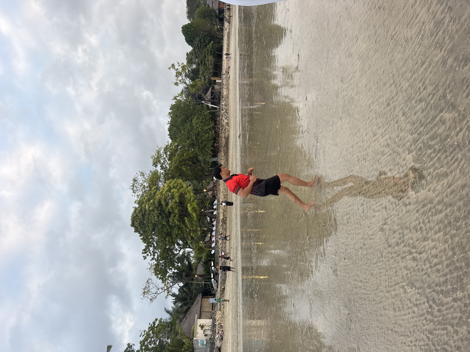 A person wearing a red shirt and black shorts is standing on a wet sandy beach. The person's body is angled towards the right, and their legs are bare. The sand is wet and reflective, with ripples and patterns visible. In the background, there is a line of trees and some buildings, suggesting a coastal setting. Other people are visible in the distance, some walking along the beach. The sky is overcast with clouds. The scene appears to be outdoors during the day, likely in a tropical location given the vegetation. The overall mood is casual and relaxed.