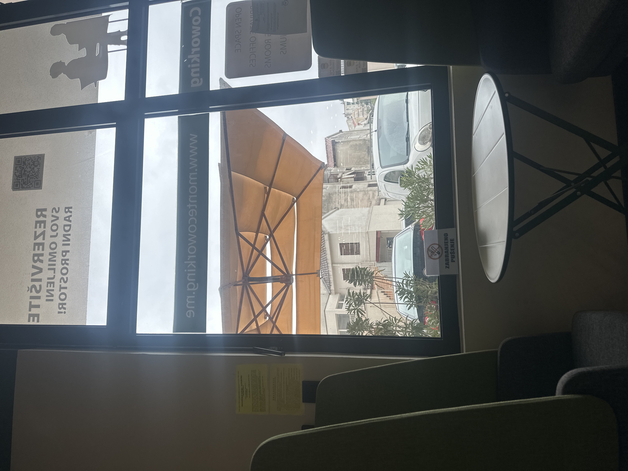 The image captures an interior view looking out a large window, likely from a coworking space or cafe, onto a street scene in Budva, Montenegro. The photograph is rotated, presenting the scene vertically.
Inside, in the immediate foreground, are several upholstered chairs in muted green and dark grey, and a light-colored, round folding table with metal legs. A light-colored wall features a yellow sheet of paper with small text, possibly instructions or information.
The prominent window is divided into several panes and features various decals and reflections. Silhouettes of people working at desks are visible on an upper-left pane. Mirrored text on the glass includes "coworking," "OPEN SPACE," "2 ROOMS," "www.montenegro," "RESERVISITE" (likely "RESERVE" or "RESERVATIONS"), and "RADIONICA" (workshop). A QR code is also present on the lower-left pane.
Outside the window, the scene is bright, indicating daytime. A large, light brown patio umbrella is a dominant feature. Beyond it, residential-style buildings with traditional red-tiled roofs and multiple windows line a street. Several cars are visible, including a white compact car resembling a Fiat 500. Green trees or bushes are scattered throughout the street view. A visible sign, facing outwards from an exterior window, clearly displays the text "ZABRANJENO PUŠENJE" (No Smoking), indicating a smoking restriction in the vicinity.
