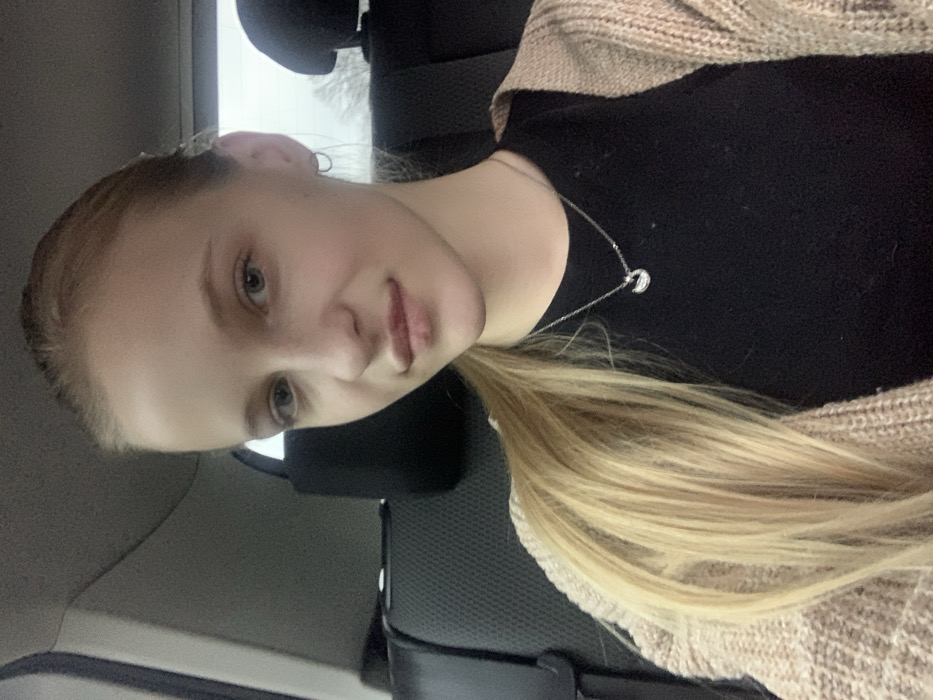 A young fair-skinned female with light blonde hair, pulled back into a ponytail, is depicted in a close-up, chest-up view, seated inside a vehicle in Oliivka, Ukraine. She has light-colored eyes and full lips, with small hoop earrings visible. She is wearing a black top beneath a light beige or brown knitted cardigan and a silver-colored necklace with a small, curved pendant. Her gaze is directed towards the camera. The background shows the dark interior of the vehicle, including parts of the rear seat and headrests. A window in the upper right reveals a blurred, overcast sky or outdoor scene.