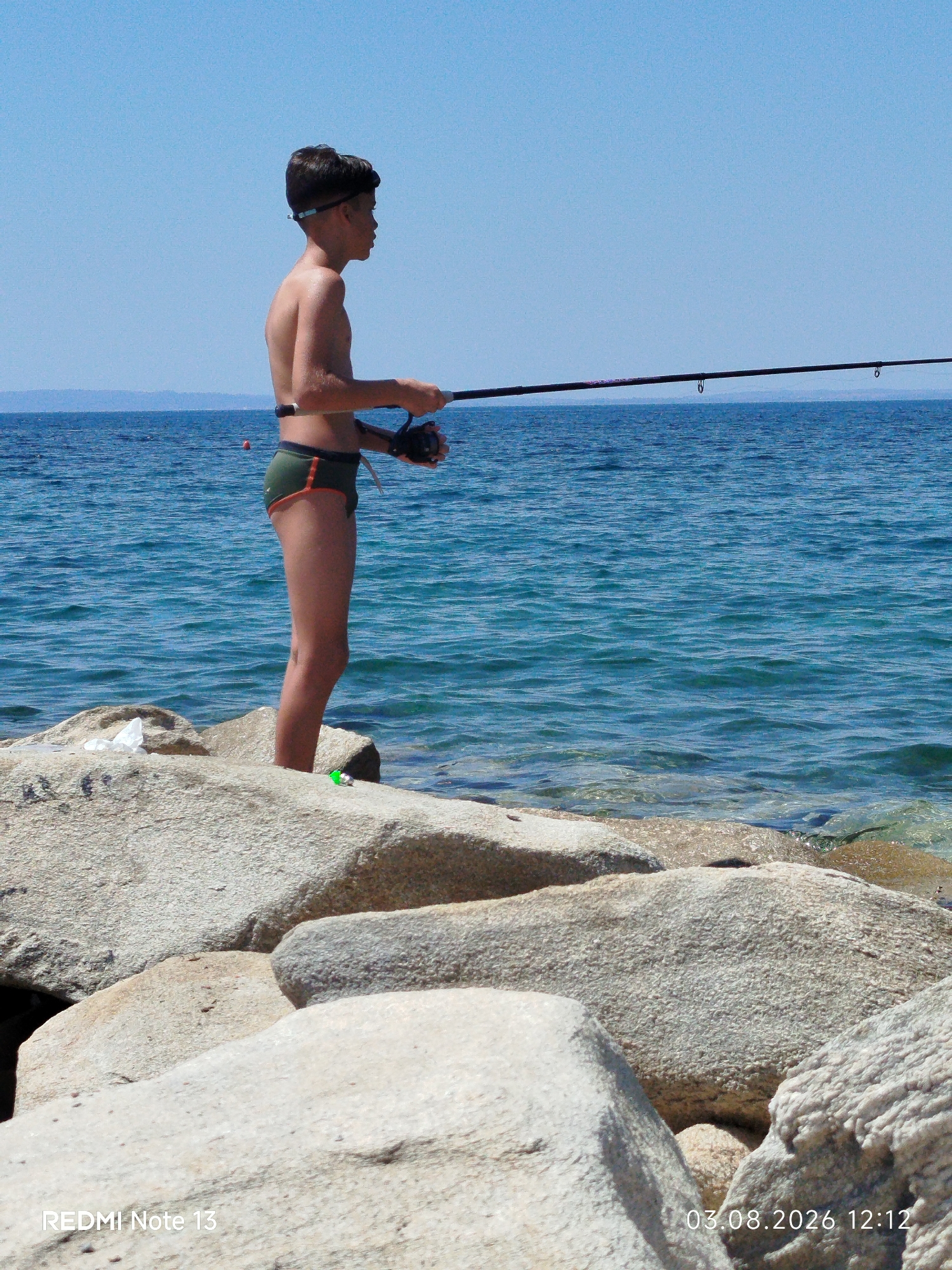 A young boy stands on a rocky shoreline, fishing in the bright sunlight. He is wearing dark green swim trunks with an orange trim and goggles pushed up onto his forehead. The boy is holding a fishing rod with a reel, casting his line into the clear blue water. The sea is calm with gentle waves lapping against the shore. In the background, the sky is a clear, vibrant blue, suggesting fair weather. The rocky terrain he stands on is composed of large, light-colored boulders.
The image appears to be taken during the day, indicated by the bright sunlight and clear sky. The location is identified as Sithonia Municipal Unit, Greece, which is a peninsula in northern Greece known for its beautiful coastlines. The overall mood is one of peaceful recreation and summer leisure.
Visible text on the image includes "REDMI Note 13" in the bottom left corner and "03.08.2026 12:12" in the bottom right corner.