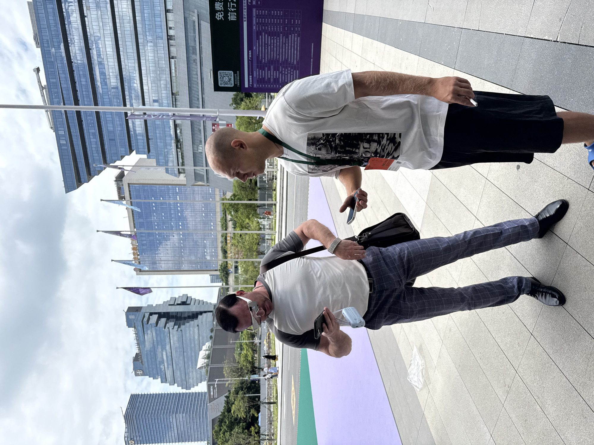 In the Haizhu District of Guangzhou, China, two men are standing on a paved walkway in front of modern glass-fronted buildings under a cloudy sky. The man on the left, wearing a white and gray t-shirt, checkered pants, and black dress shoes, is looking down at his smartphone, holding a small white and blue device. He has sunglasses on and a bag slung over his shoulder. The man on the right, in a white t-shirt, black shorts, and a green lanyard with a red tag, is also holding a smartphone and appears to be smoking a cigarette. A large purple sign with Chinese characters is visible behind them.