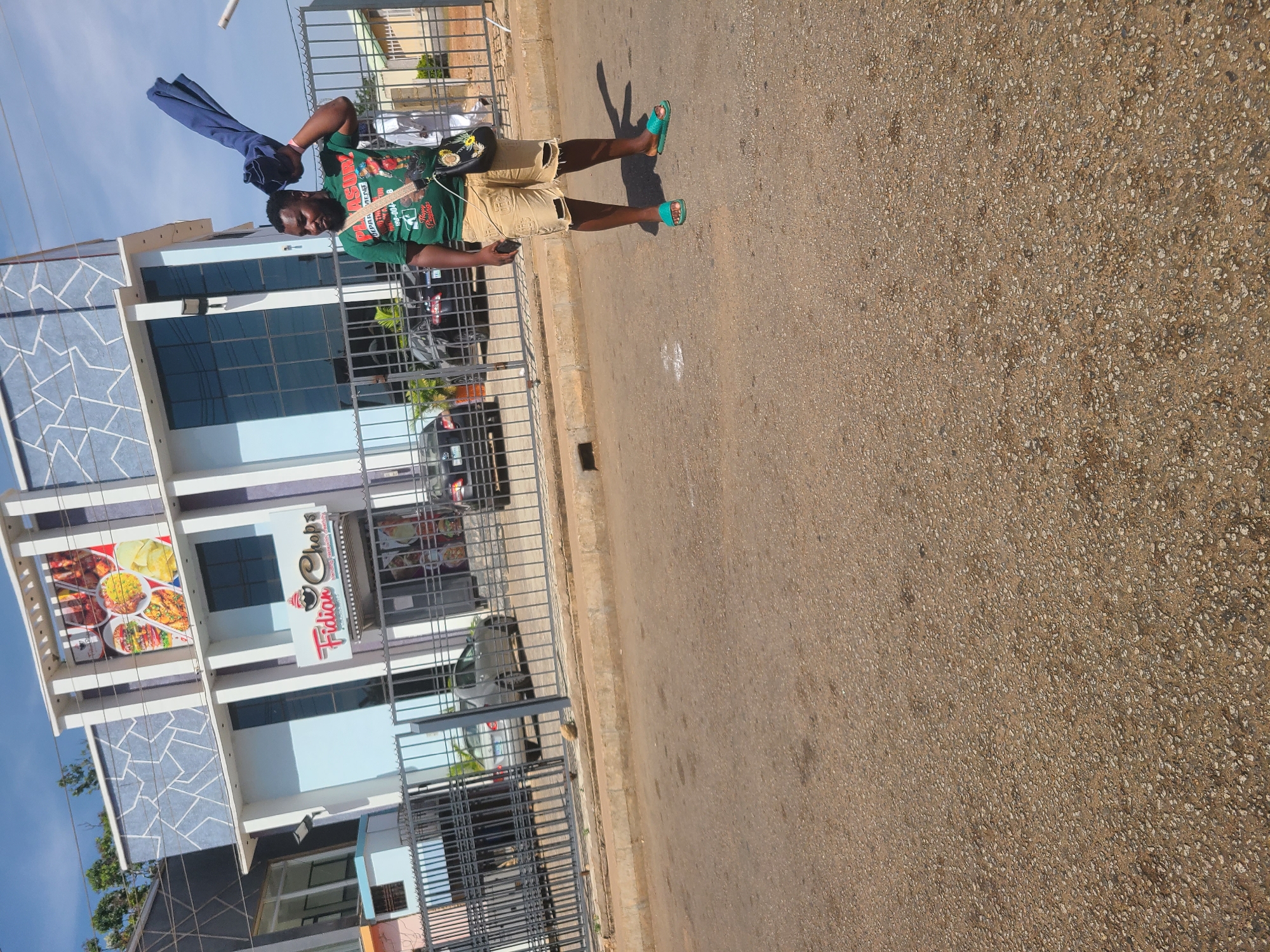 A street-level photograph depicts a man standing on a paved street beside a building. The man is wearing a green graphic t-shirt, khaki shorts, and light-blue slide sandals. He holds a blue object, possibly a garment, aloft in his left hand and appears to be looking at a mobile phone in his right hand. A dark cross-body bag is slung across his torso.
Behind the man is a building with a multi-story facade, composed of blue and white panels. A sign reading "Fidian Chopps" is prominently displayed on the building's second story. Metal security bars are visible along the sidewalk in front of the building.
The street stretches horizontally across the frame, with a shadow extending from the man onto the pavement. The pavement appears to be tan in color and shows signs of wear.
Based on the signage and architectural style, this scene is most likely taken in Jos, Nigeria.