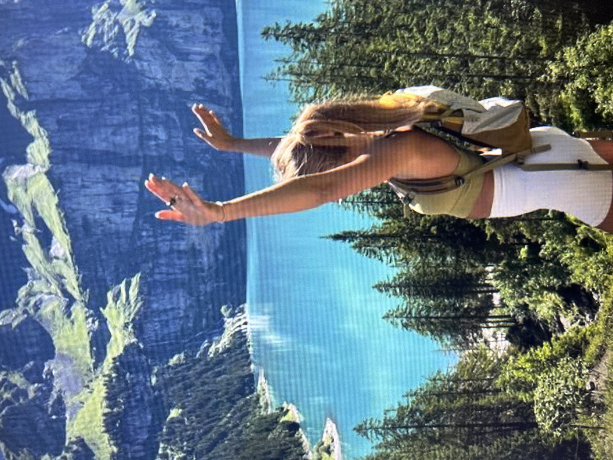 A woman with blonde hair in a ponytail, wearing a green crop top, white shorts, and a backpack, stands with her arms raised against a backdrop of a stunning turquoise lake and dramatic mountains. The mountains are steep and rocky, with patches of green vegetation and dark green pine trees covering their lower slopes. The lake is calm and reflects the sky and surrounding landscape. Pine trees are also abundant in the foreground and on the hillsides. The lighting suggests it is daytime and sunny. The scene appears to be a natural, mountainous landscape, likely a hiking or scenic viewpoint. There is no indication of Qarağandı city administration, Kazakhstan in this image.