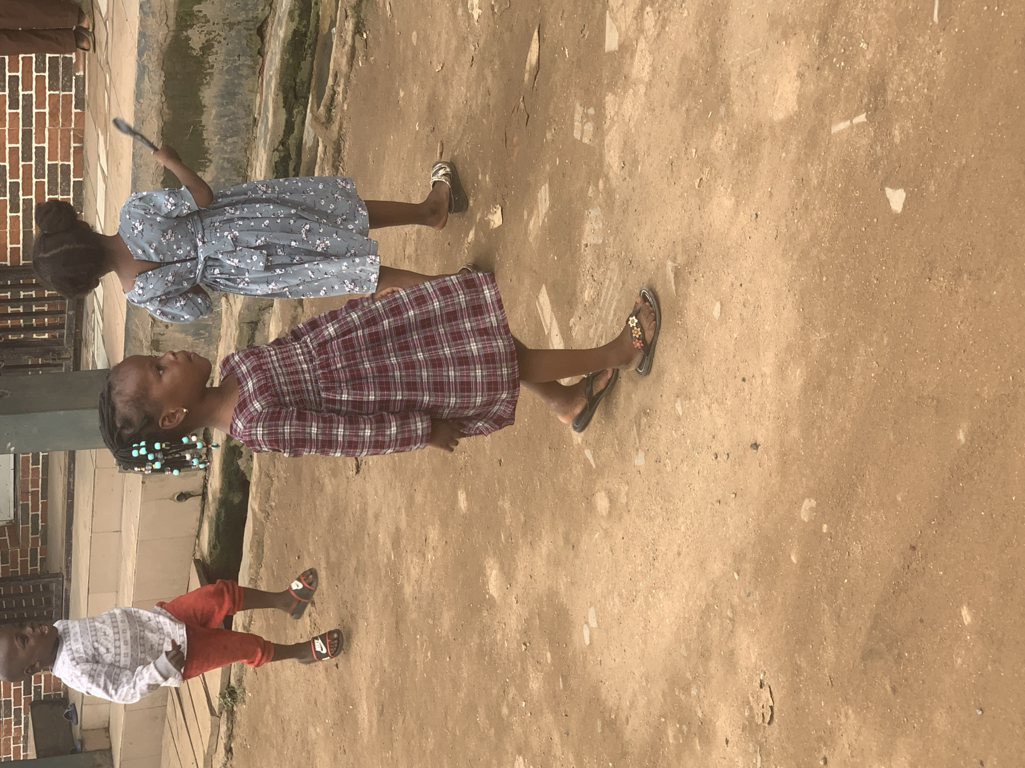Three children, two girls and one boy, are walking on a dirt path in an urban setting in Shagamu, Nigeria. The boy, in the foreground on the left, wears a white, long-sleeved shirt with a grey pattern and red pants. He has on black slide sandals. The girl in the center wears a red and white plaid dress and black sandals decorated with flowers. Her hair is styled in braids with blue beads. The second girl, on the right, is further back and wears a blue floral-print dress and sandals. She is holding a dark-colored object, possibly a utensil, in her right hand. The background shows a brick building and concrete stairs. The ground is composed of reddish-brown dirt.