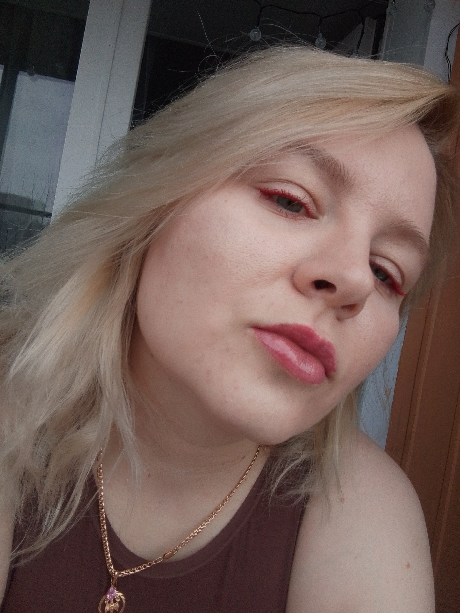 A fair-skinned person with blonde hair and a reddish lip color is shown in a close-up, selfie-style shot. The person is wearing red eyeliner. A gold necklace with a pendant hangs around the person's neck. The person is wearing a brown top. Behind the person is a window frame and door, suggesting an indoor location. The location in the city of Izhevsk, Russia, cannot be determined from this image.